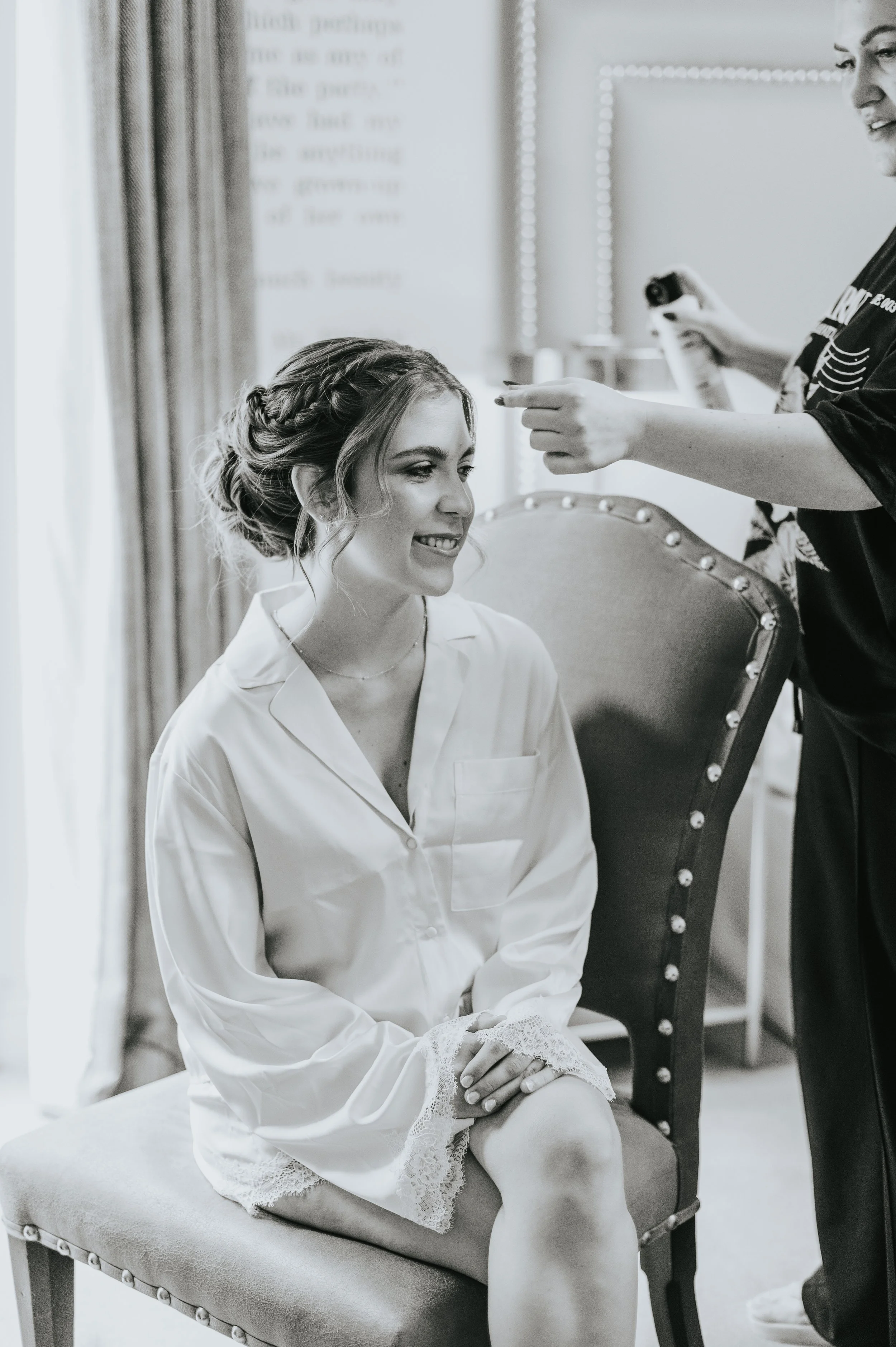 A woman getting her hair styled, sitting on a chair, smiling and looking to the side, as a hairstylist applies finishing touches to her hair in a well-lit room.