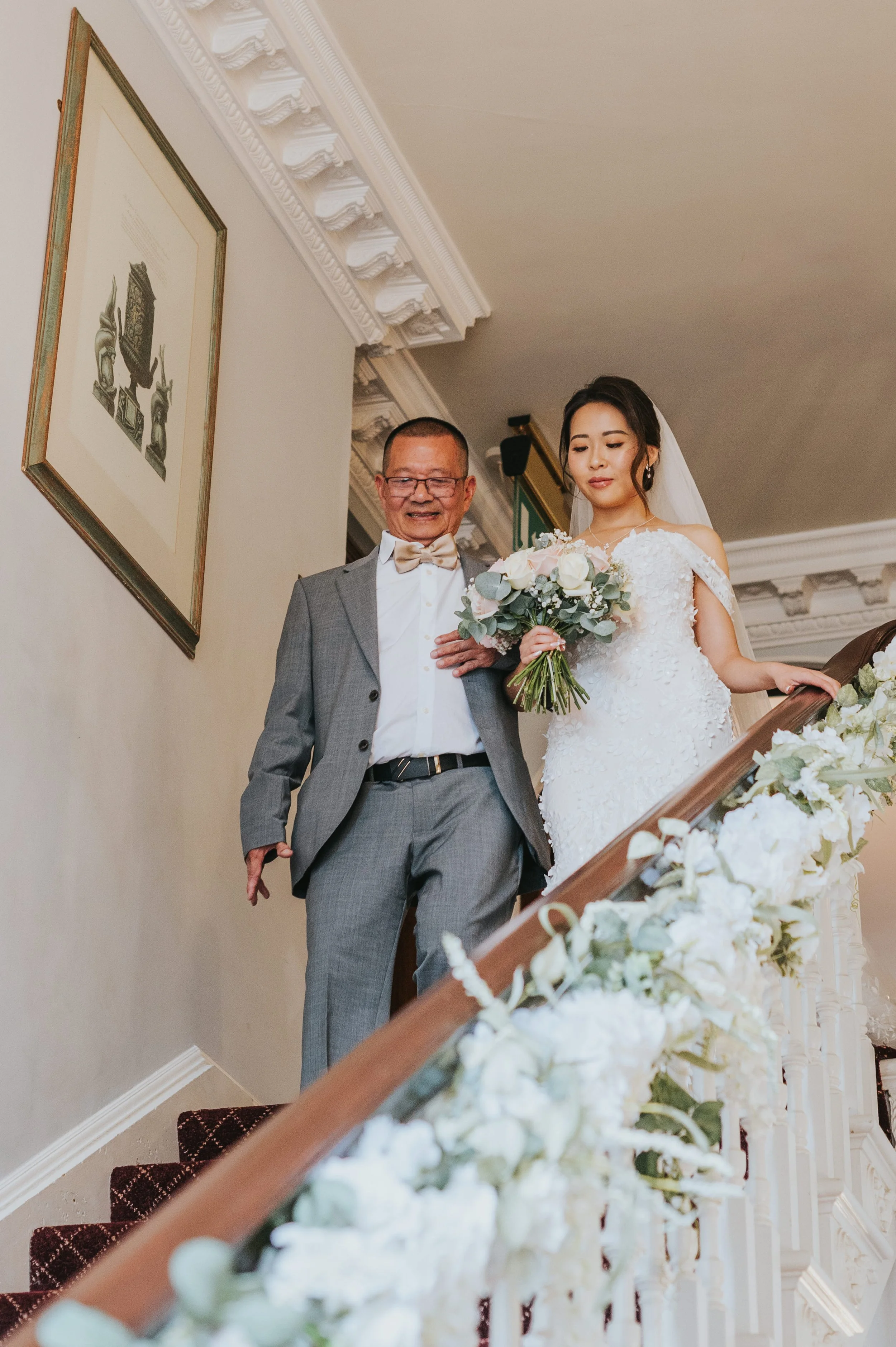 A bride in a white wedding dress holding a bouquet, descending a staircase with a man in a gray suit, white shirt, and bow tie, surrounded by white floral decorations on the staircase railing.