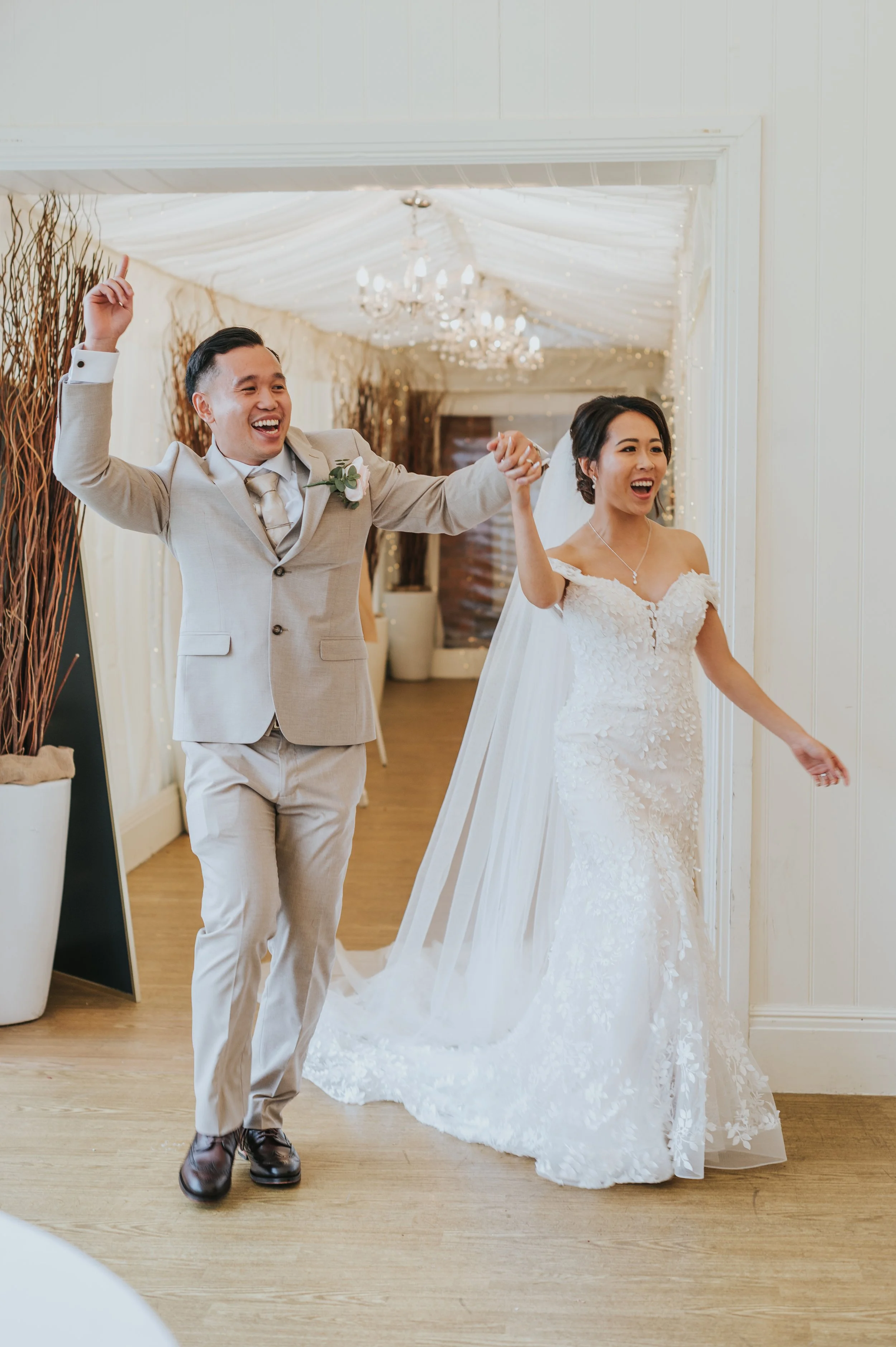 Newlywed couple celebrating indoors, the groom in a beige suit and the bride in a white lace wedding gown, holding hands and smiling. Rebecca Louise Photography Essex Wedding photographer Quendon Hall