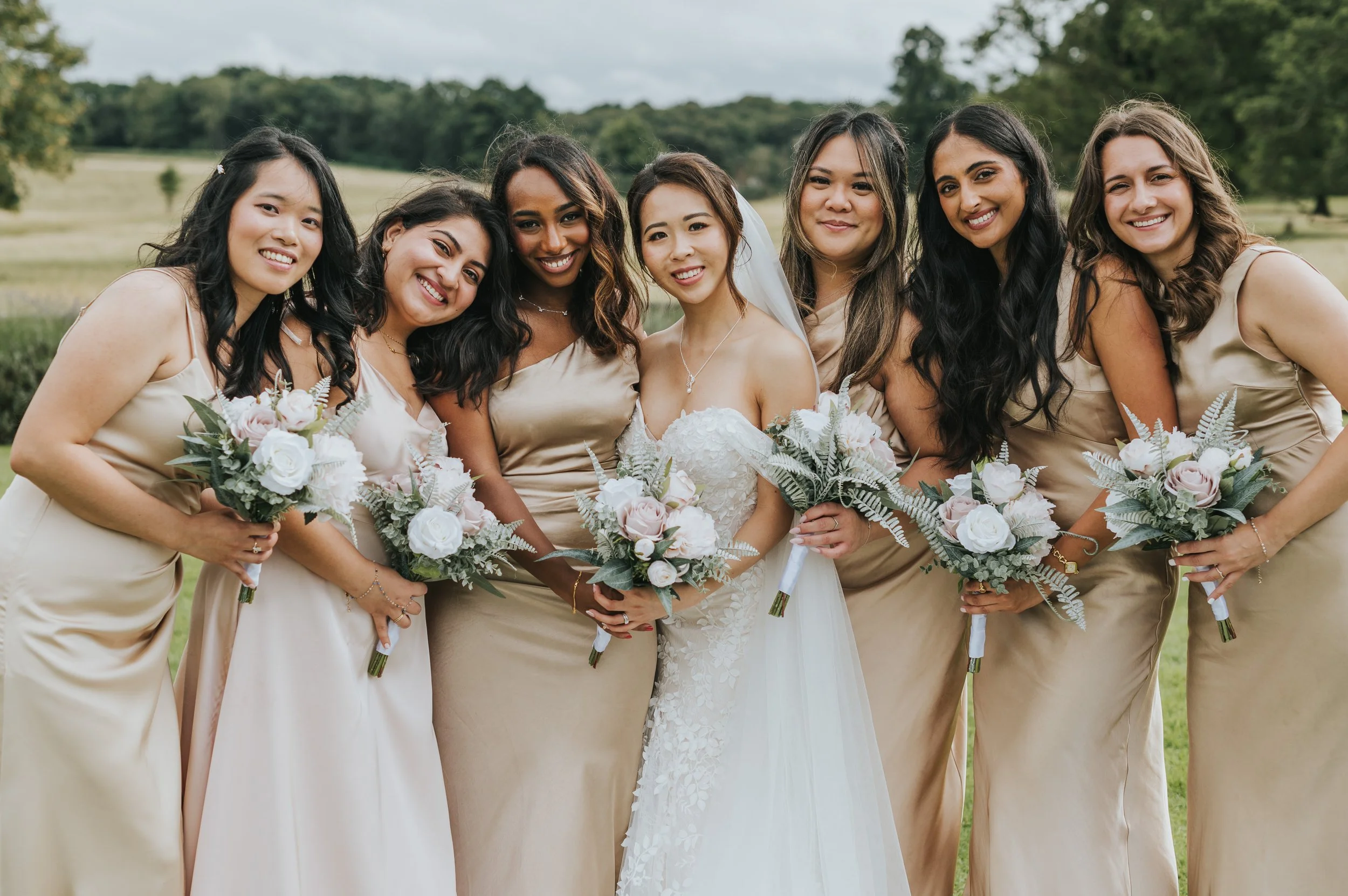 Group of eight women at a wedding outdoors, dressed in beige dresses, holding bouquets, standing together smiling. Rebecca Louise Photography Essex Wedding photographer