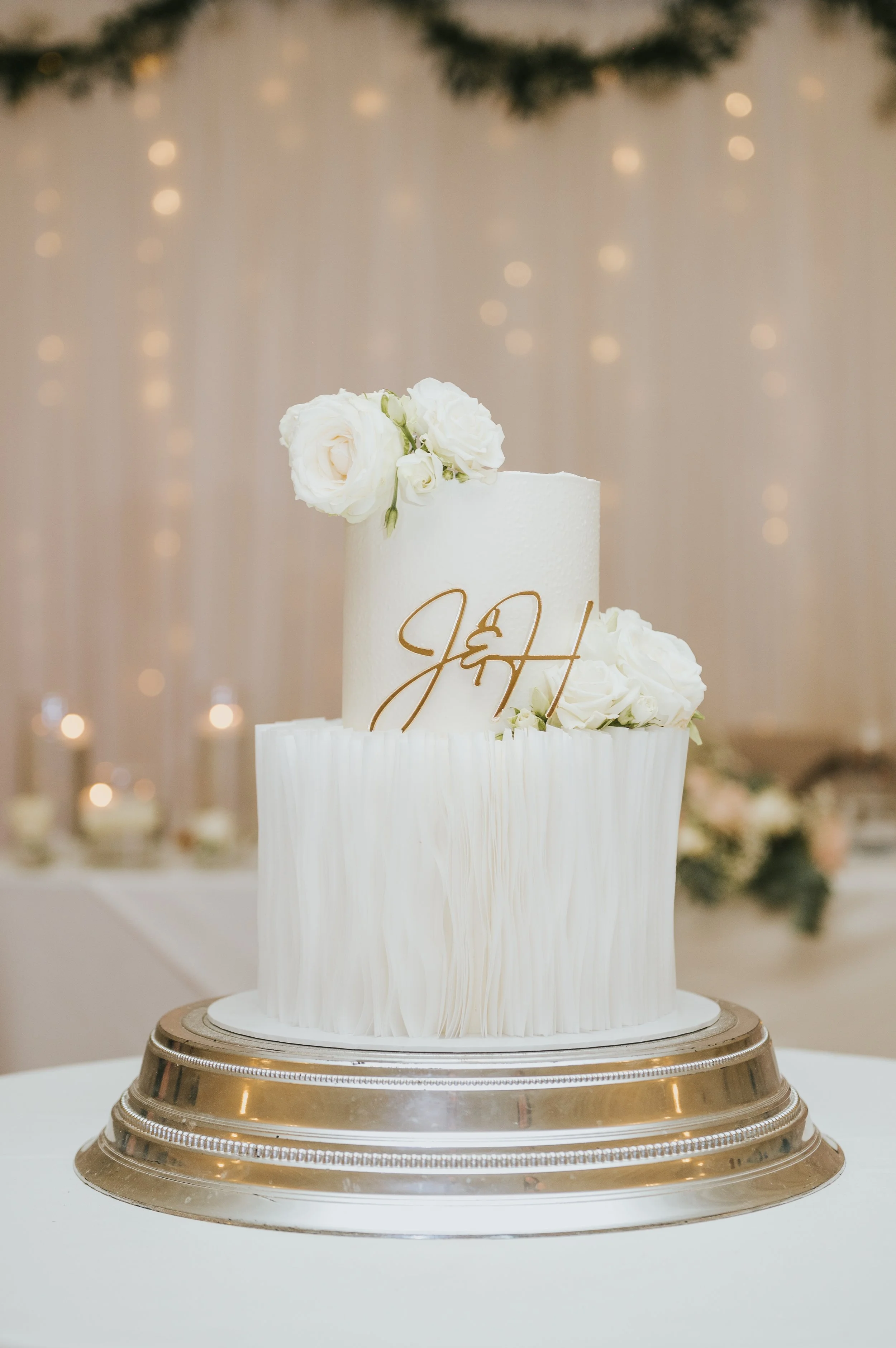 Elegant white wedding cake with floral decorations and gold cursive initials 'J & H' on top, displayed on a silver cake stand.