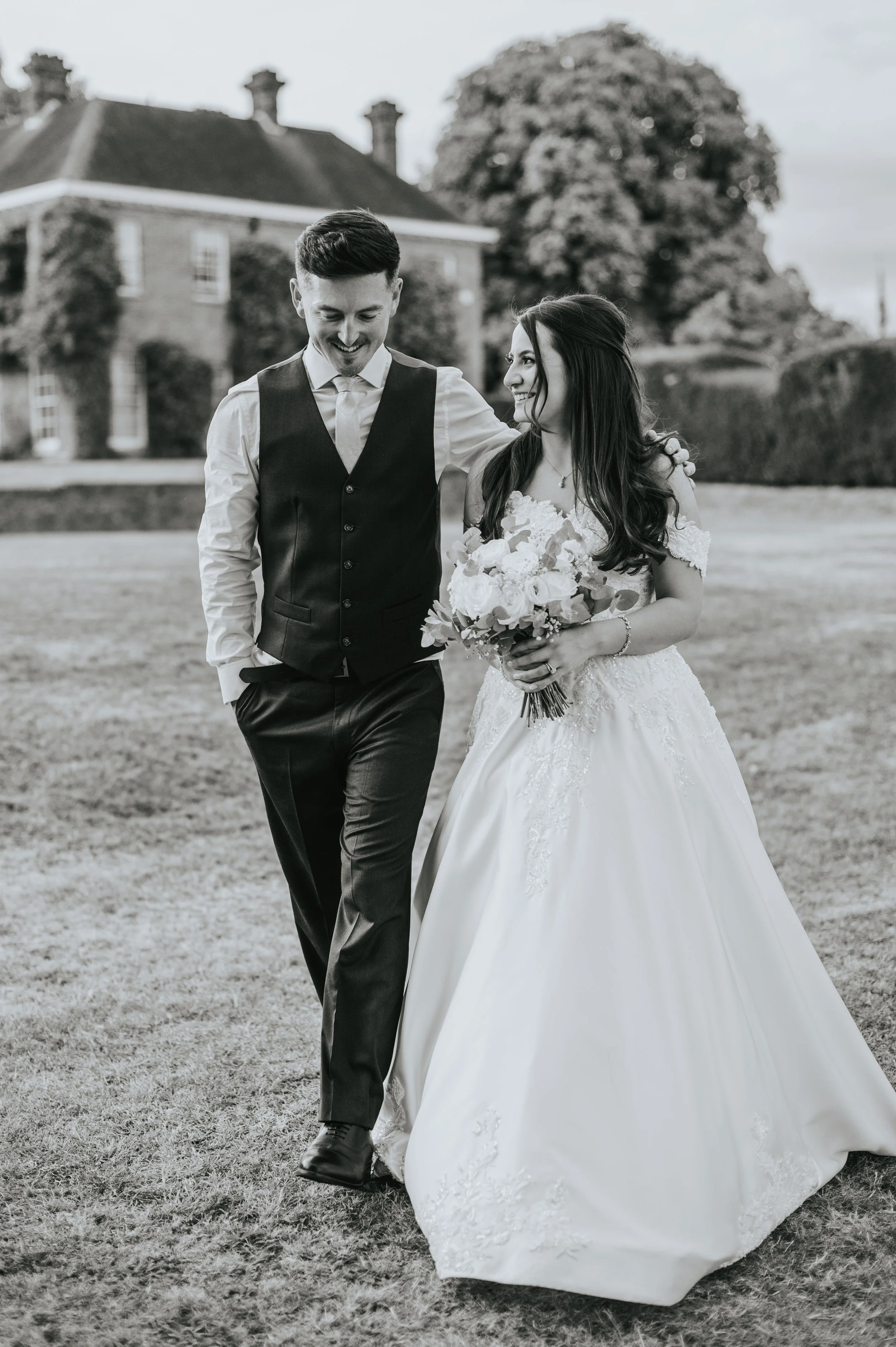 Black and white photo of a bride and groom walking on grass, smiling at each other, with a large house and trees in the background. The bride is holding a bouquet of flowers and wearing a wedding gown, while the groom is dressed in a vest and dress s
