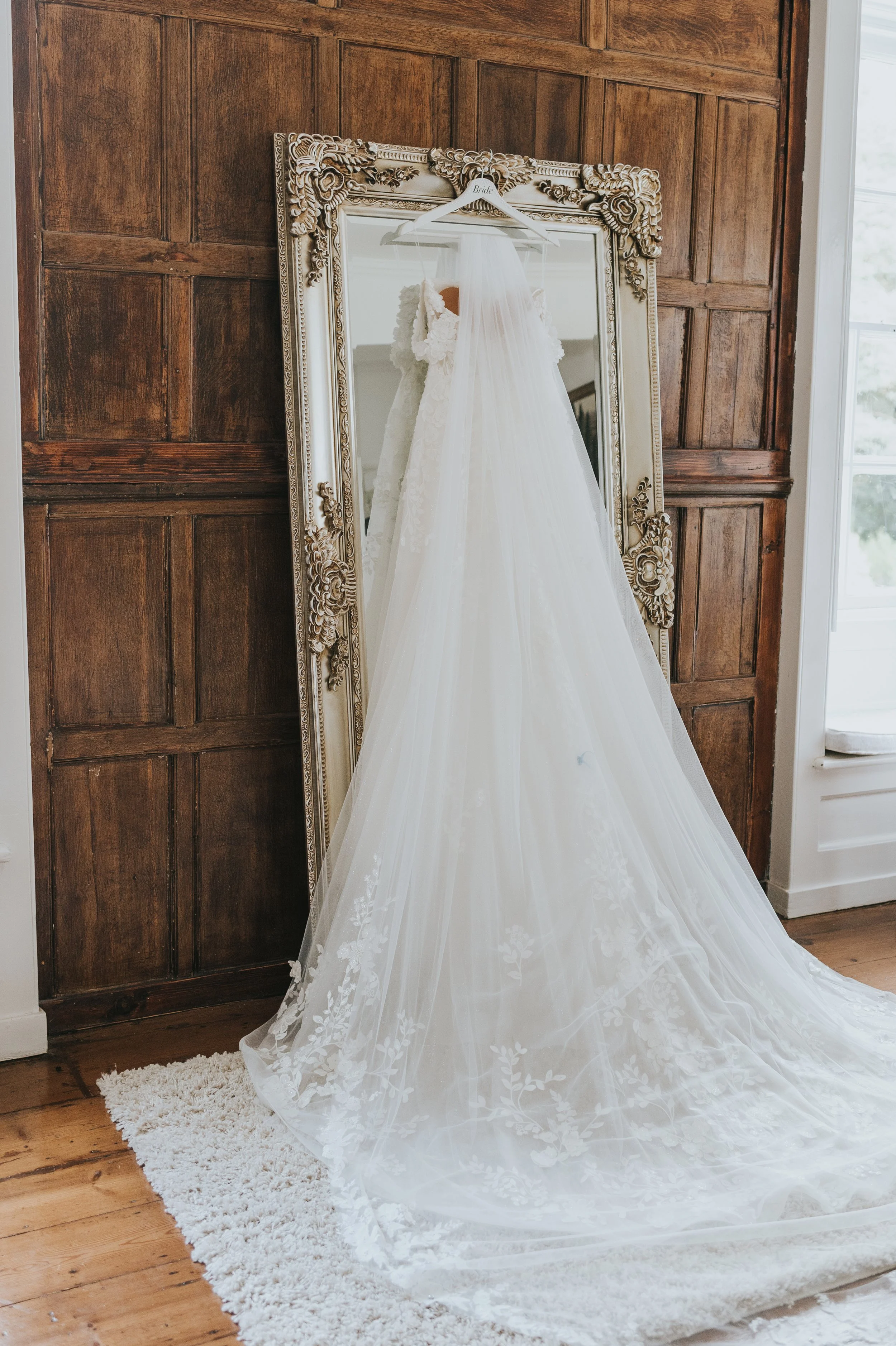 Wedding dress hanging on a hanger labeled Bride, reflected in a large ornate mirror, with a wooden wall and a window in the background