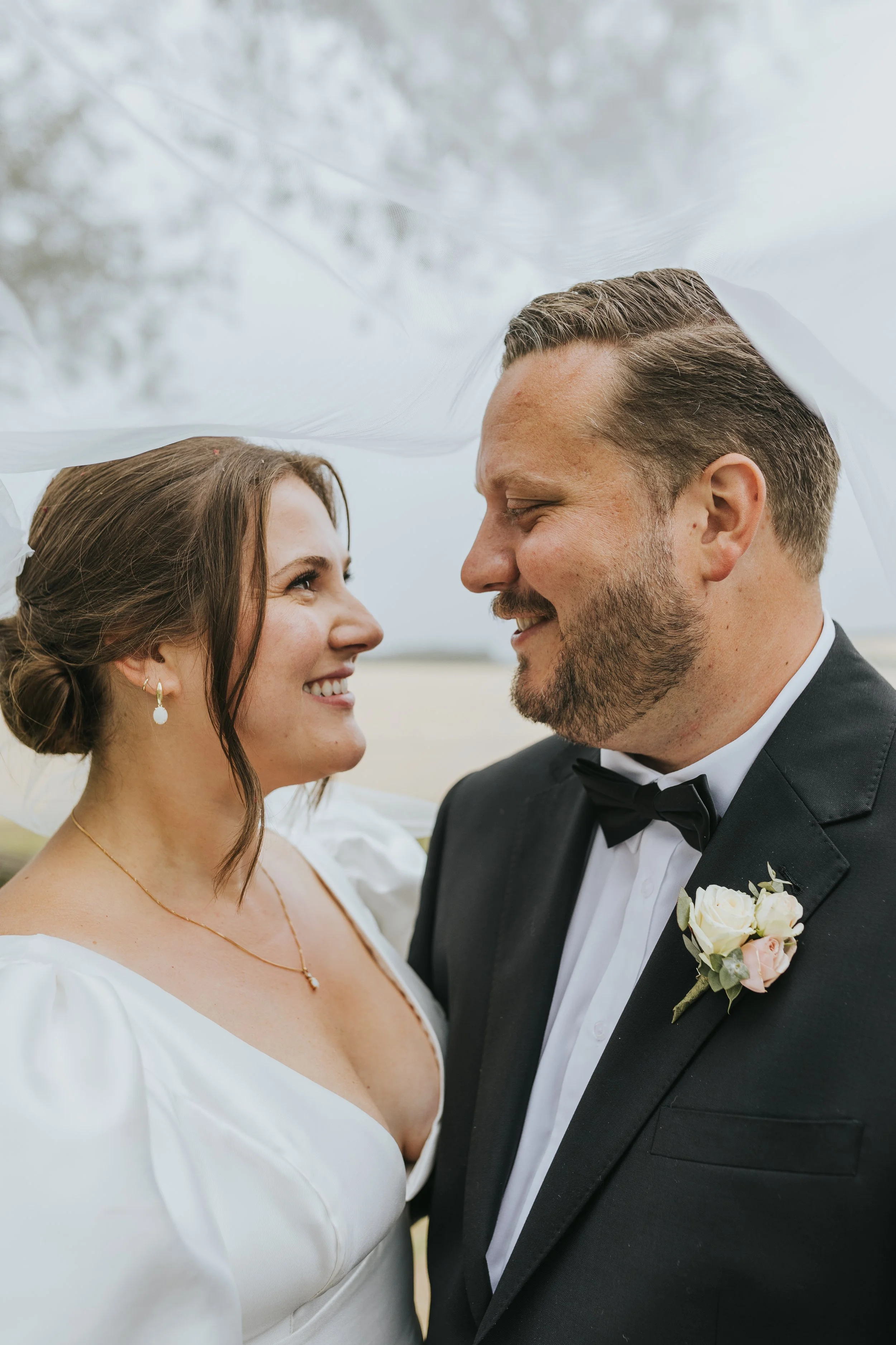 A bride and groom smiling at each other under a veil outdoors on their wedding day.