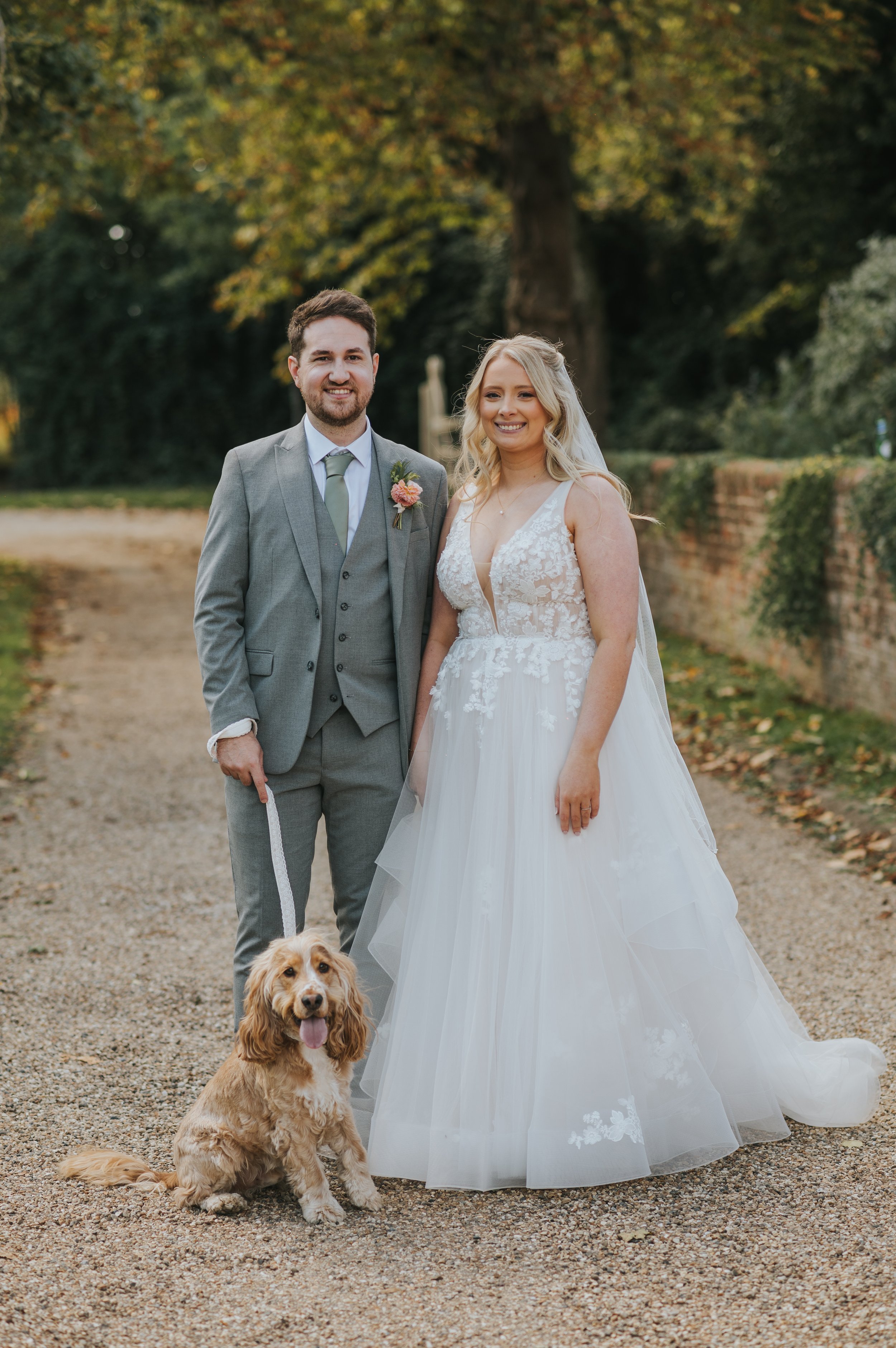 A bride and groom standing outdoors on a gravel path, with a dog sitting in front of them. The groom is in a gray suit with a boutonniere, and the bride is in a white wedding gown with floral lace detail. Rebecca Louise Photography Essex Wedding 
