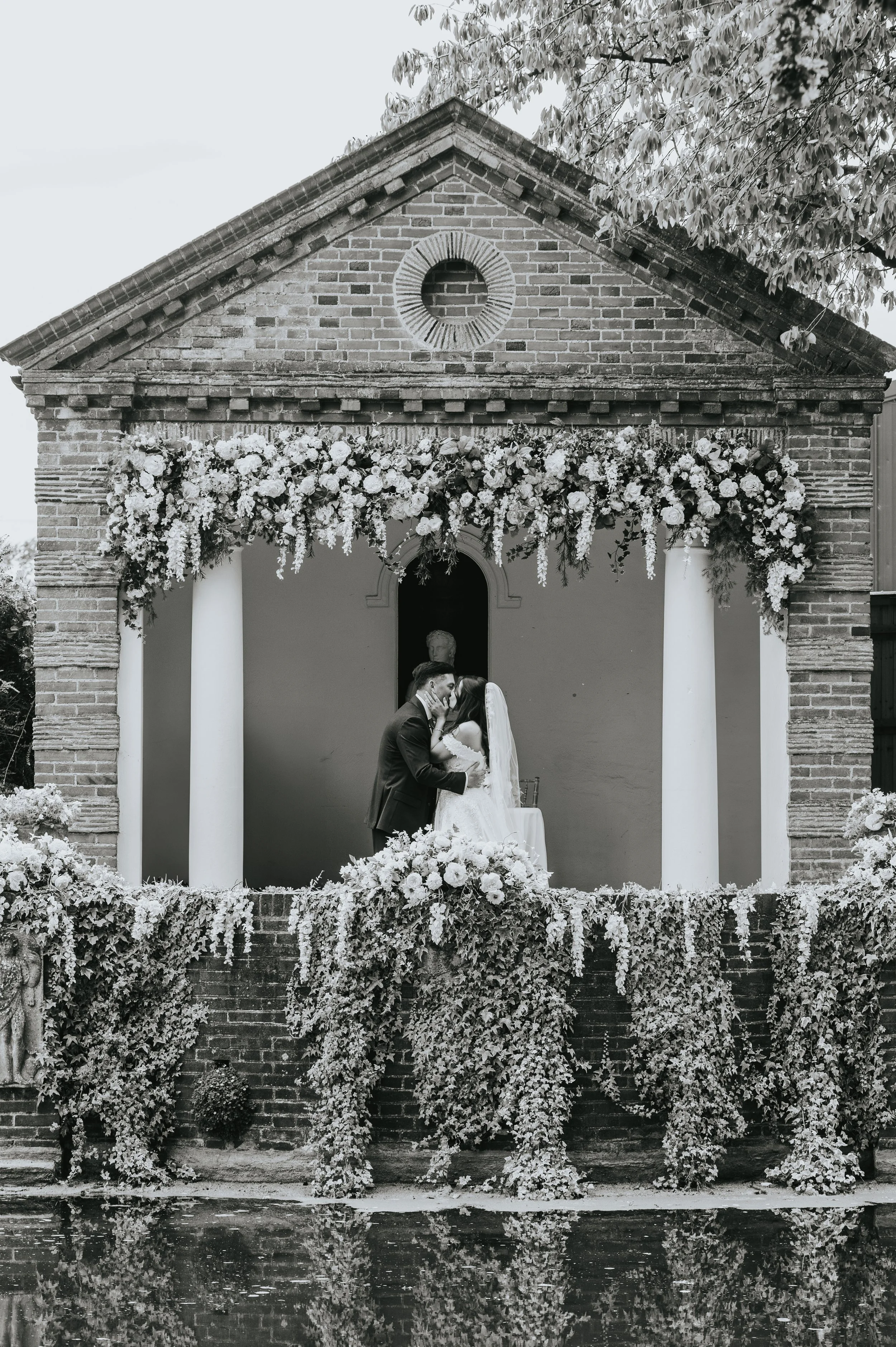 A black and white photo of a wedding ceremony taking place on a small outdoor stage decorated with lush floral arrangements. Rebecca Louise Photography Essex Wedding photographer. Micklefield Hall