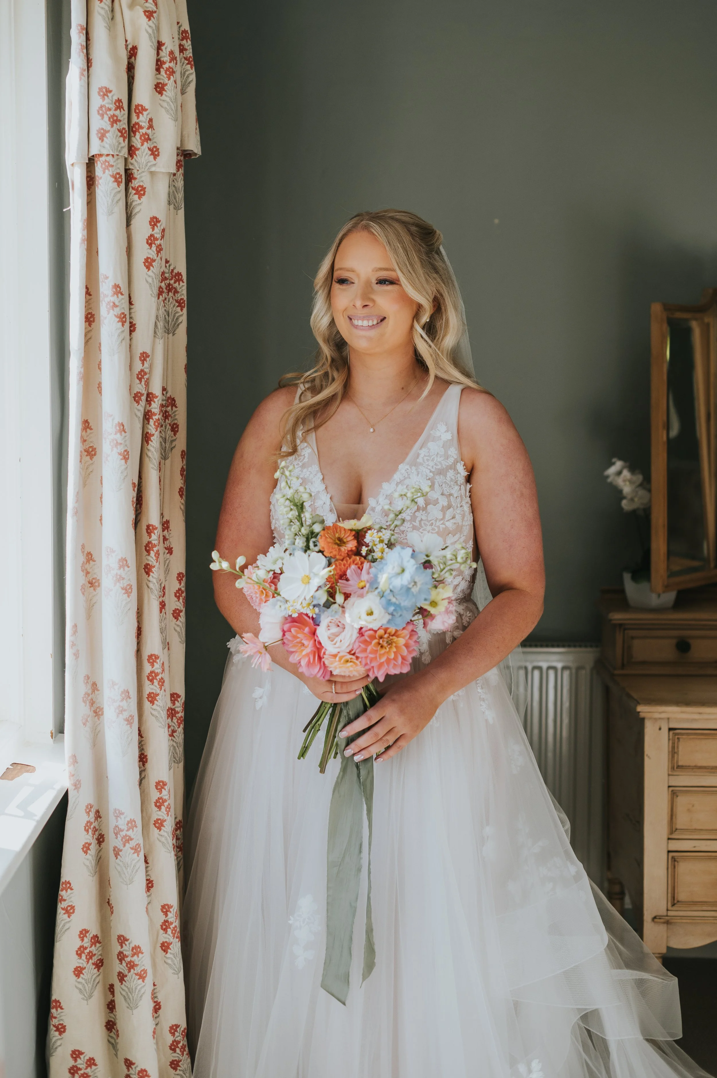 A bride in a white wedding dress holding a colorful bouquet of flowers, standing indoors near a window with floral curtains and a wooden dresser in the background.