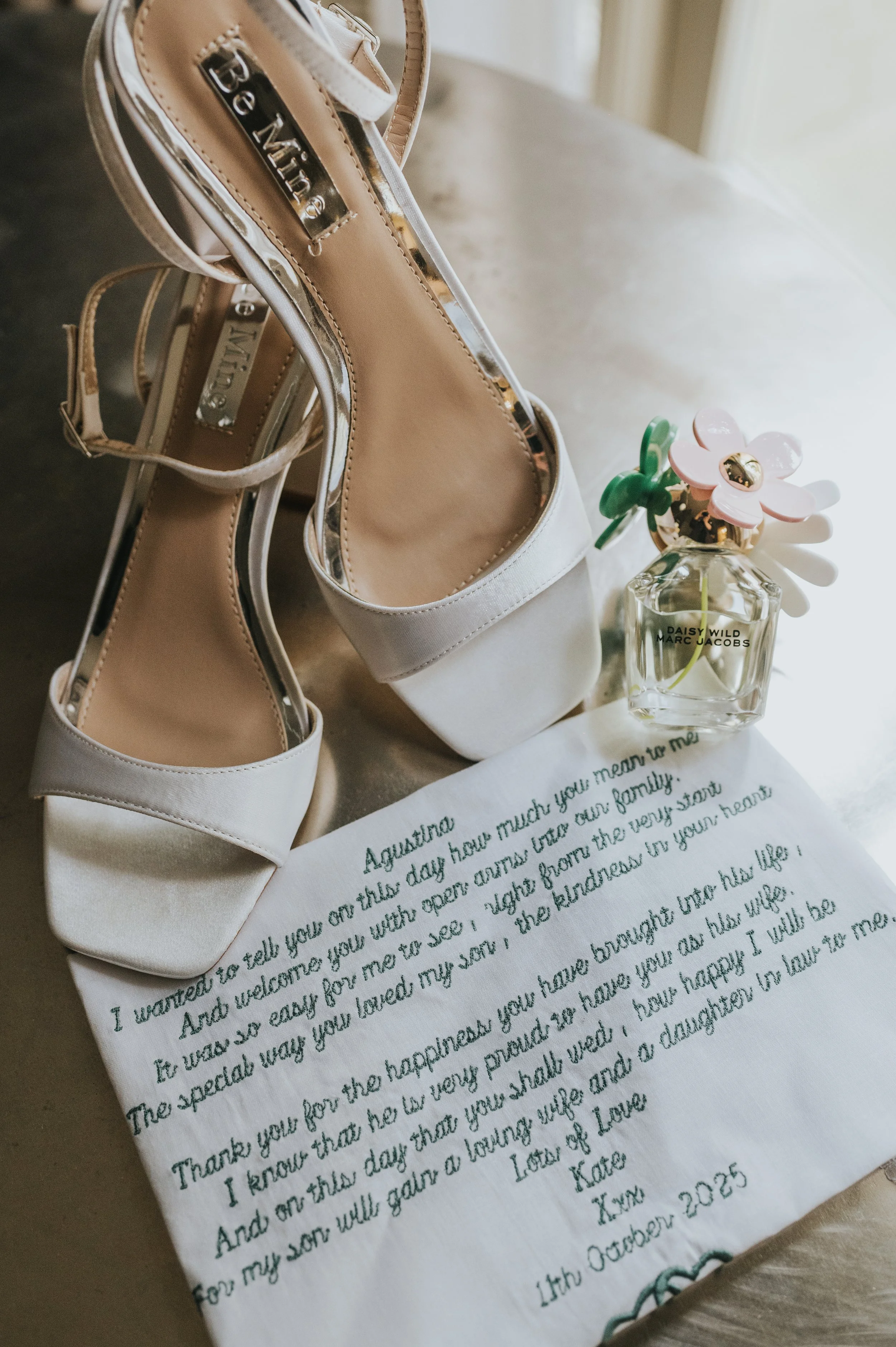 A pair of white high-heeled shoes, a glass perfume bottle with pink and green flower-shaped cap, and a handwritten note with a heartfelt message, all arranged on a light-colored fabric surface. Rebecca Louise Photography Essex Wedding photographer