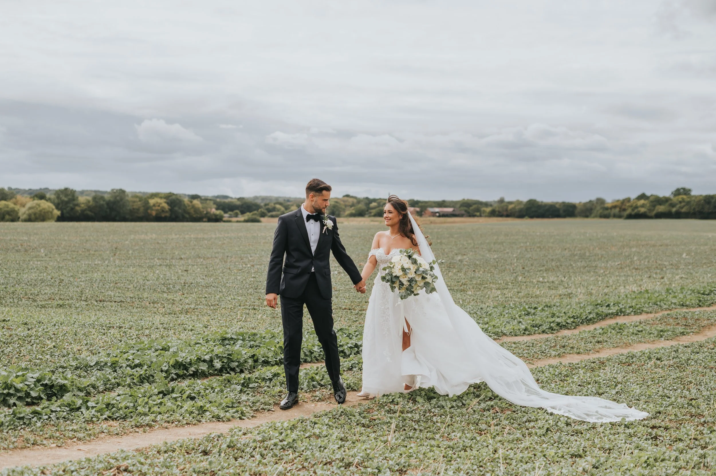 A bride and groom holding hands and walking in a field on their wedding day. The bride is wearing a white off-the-shoulder wedding gown with a veil, and carrying a large bouquet of white flowers. The groom is dressed in a black tuxedo with a bow tie.