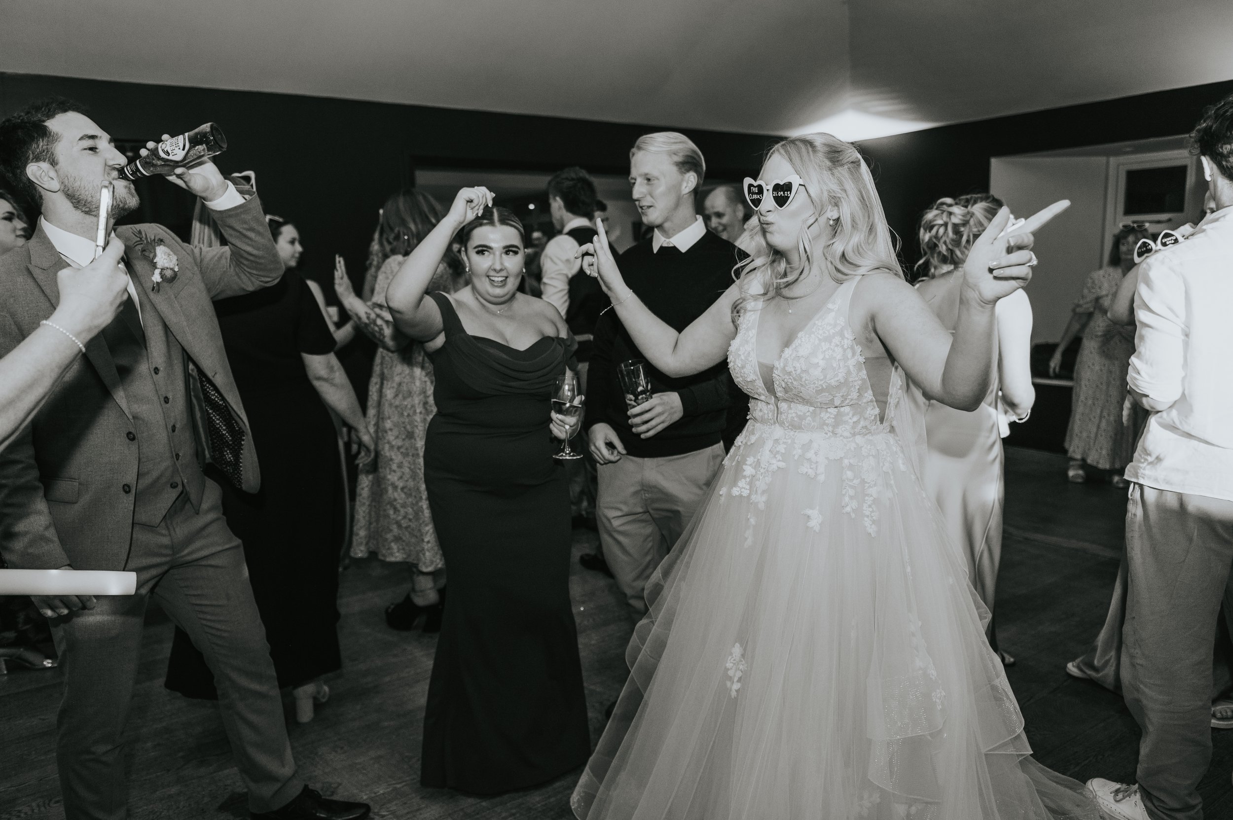 People dancing and socializing at a wedding reception, with the bride in a wedding gown and guests holding drinks. Rebecca Louise Photography Essex Wedding photographer Houchins