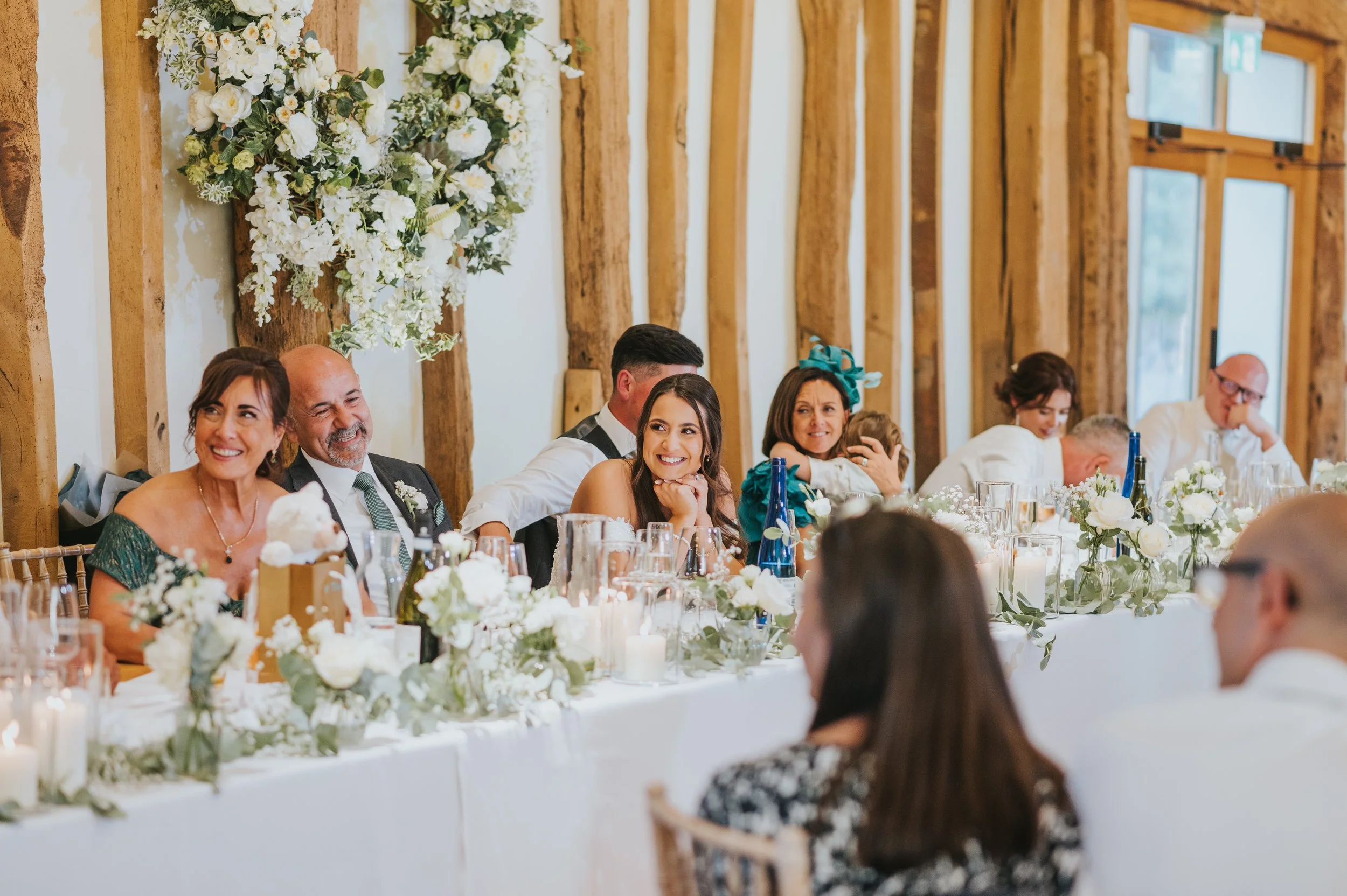 People sitting at a decorated wedding reception table, smiling and enjoying the moment, with floral arrangements, candles, and wine bottles.