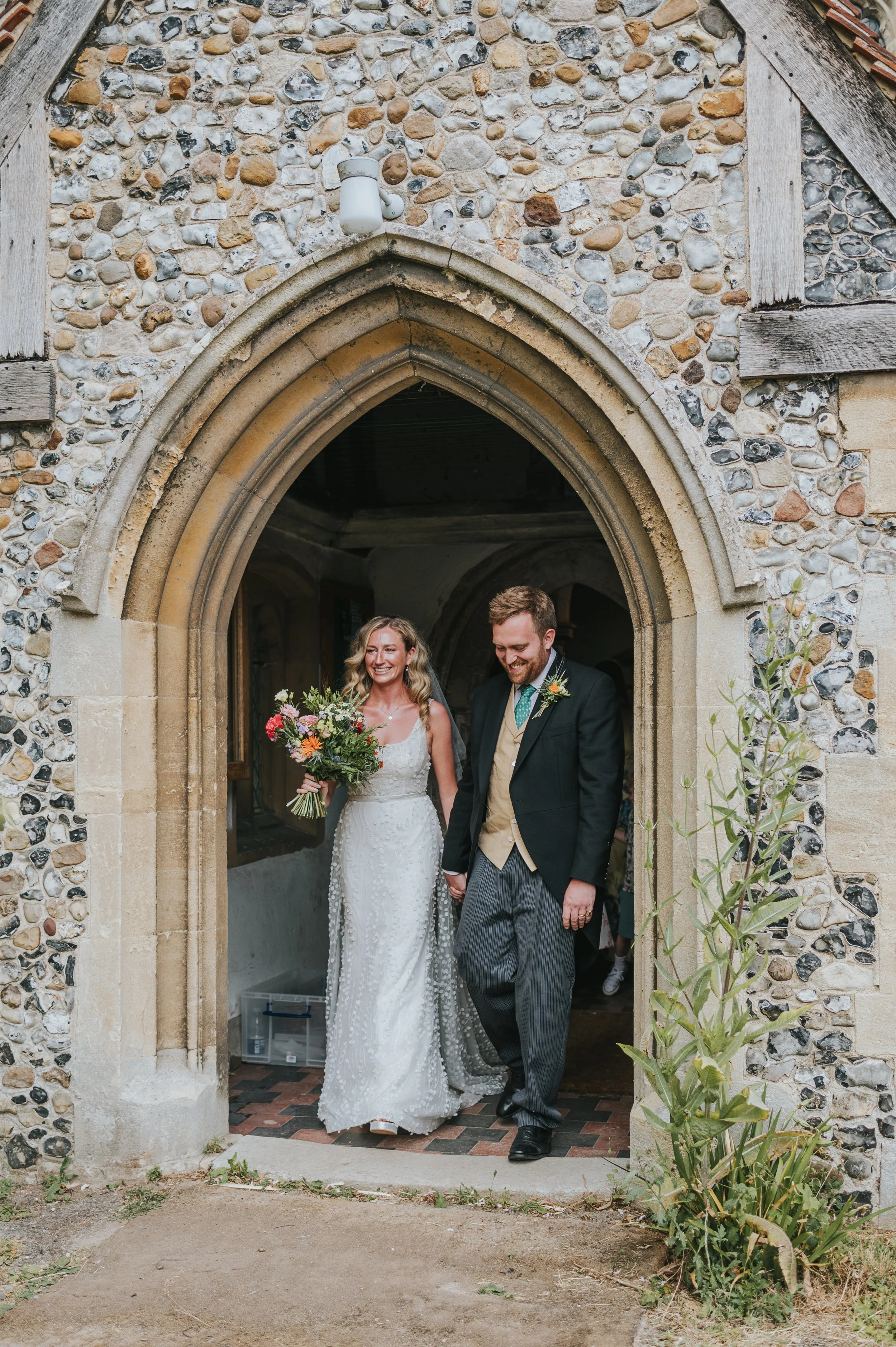 A bride and groom walking out of a stone church, holding hands and smiling, with the bride holding a bouquet of colorful flowers.
