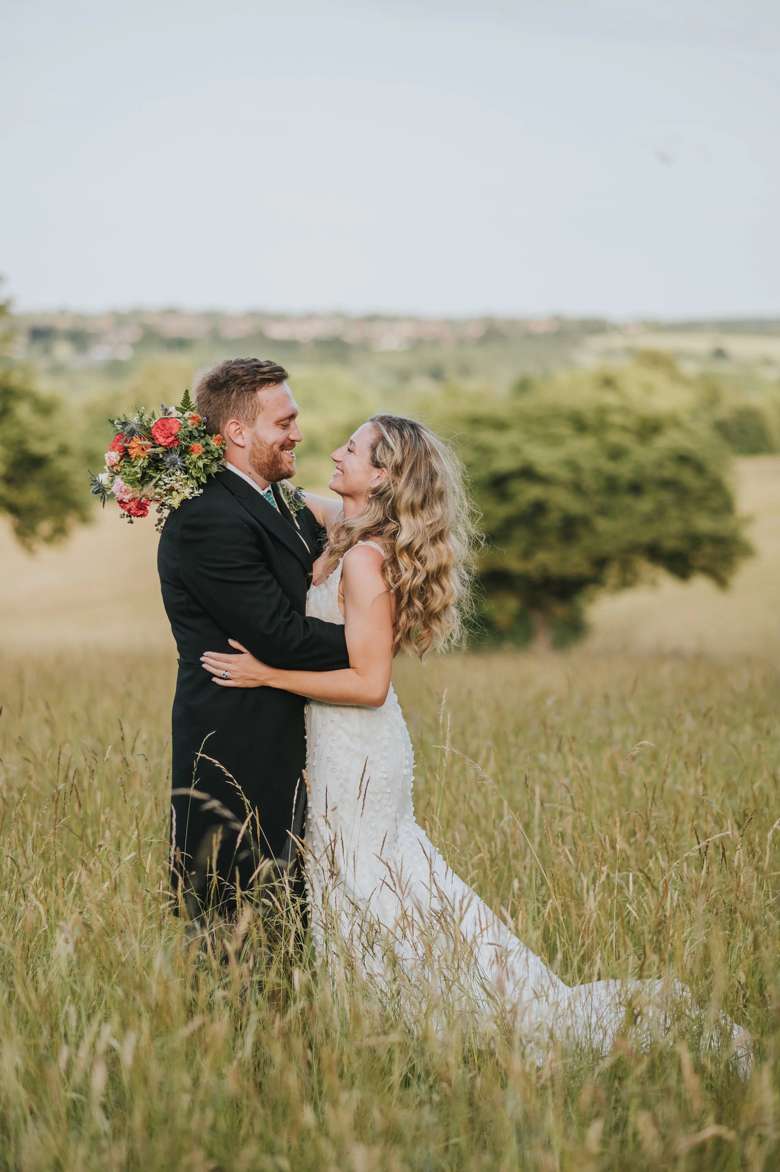 A bride and groom embrace in a grassy field with trees and a distant landscape, smiling at each other on their wedding day.