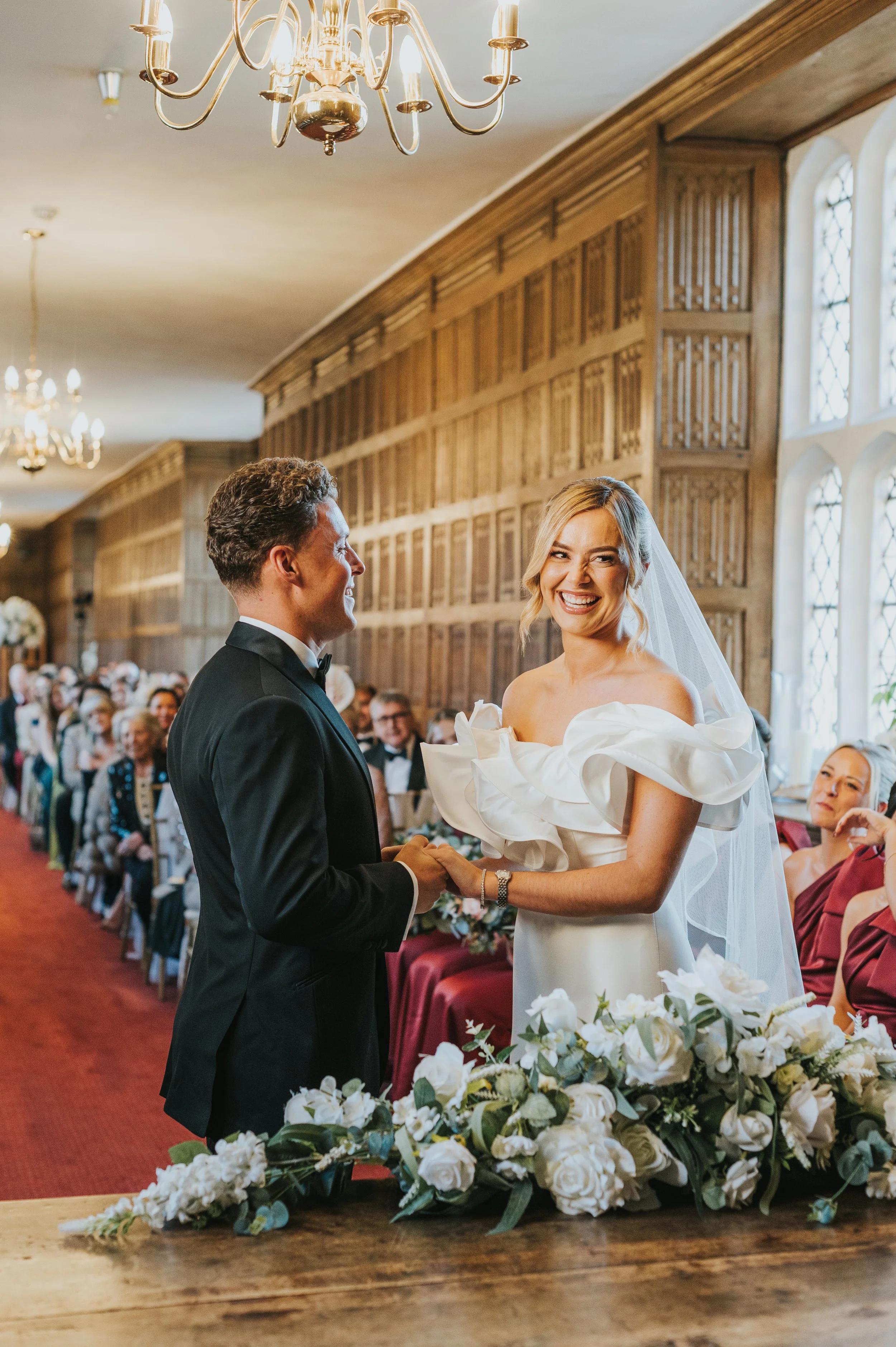 Bride and groom holding hands during wedding ceremony inside a wood-paneled room with large windows and chandeliers, surrounded by seated guests.