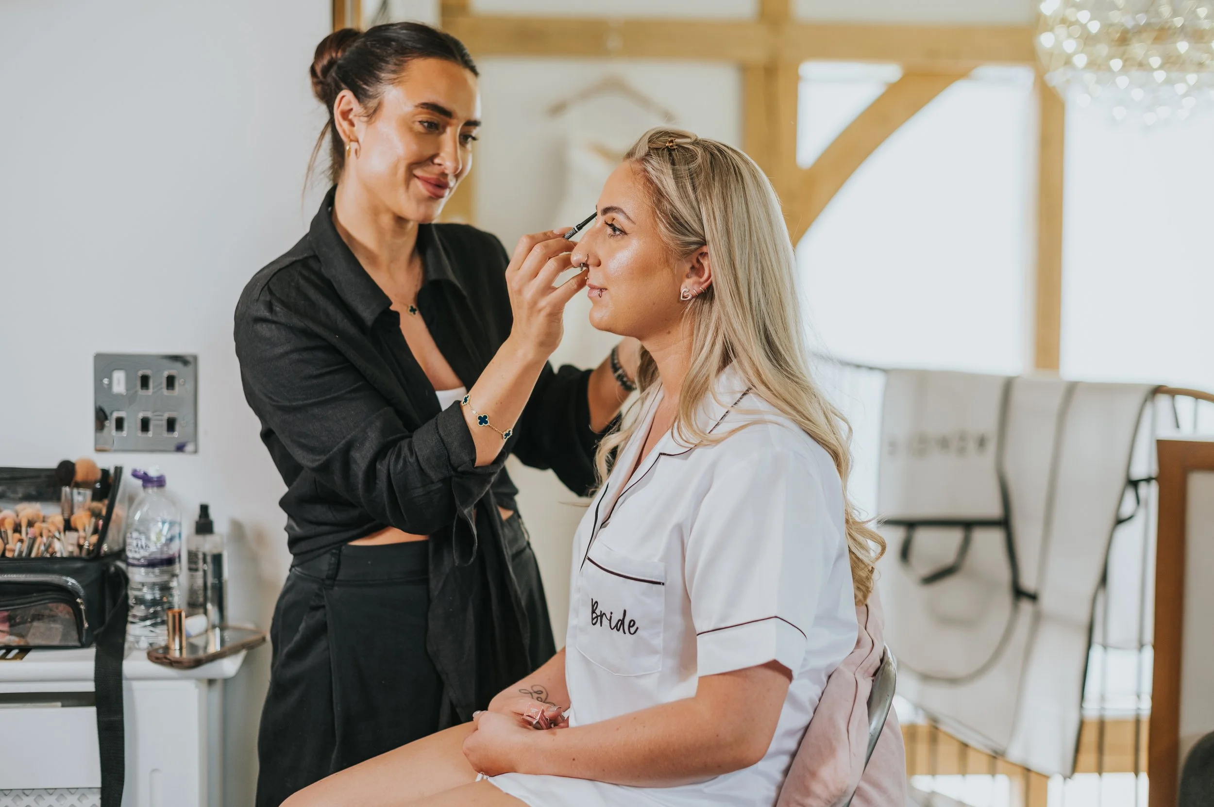A makeup artist applying makeup to a bride's face in a bright room with wooden beams, with makeup tools on a nearby table.