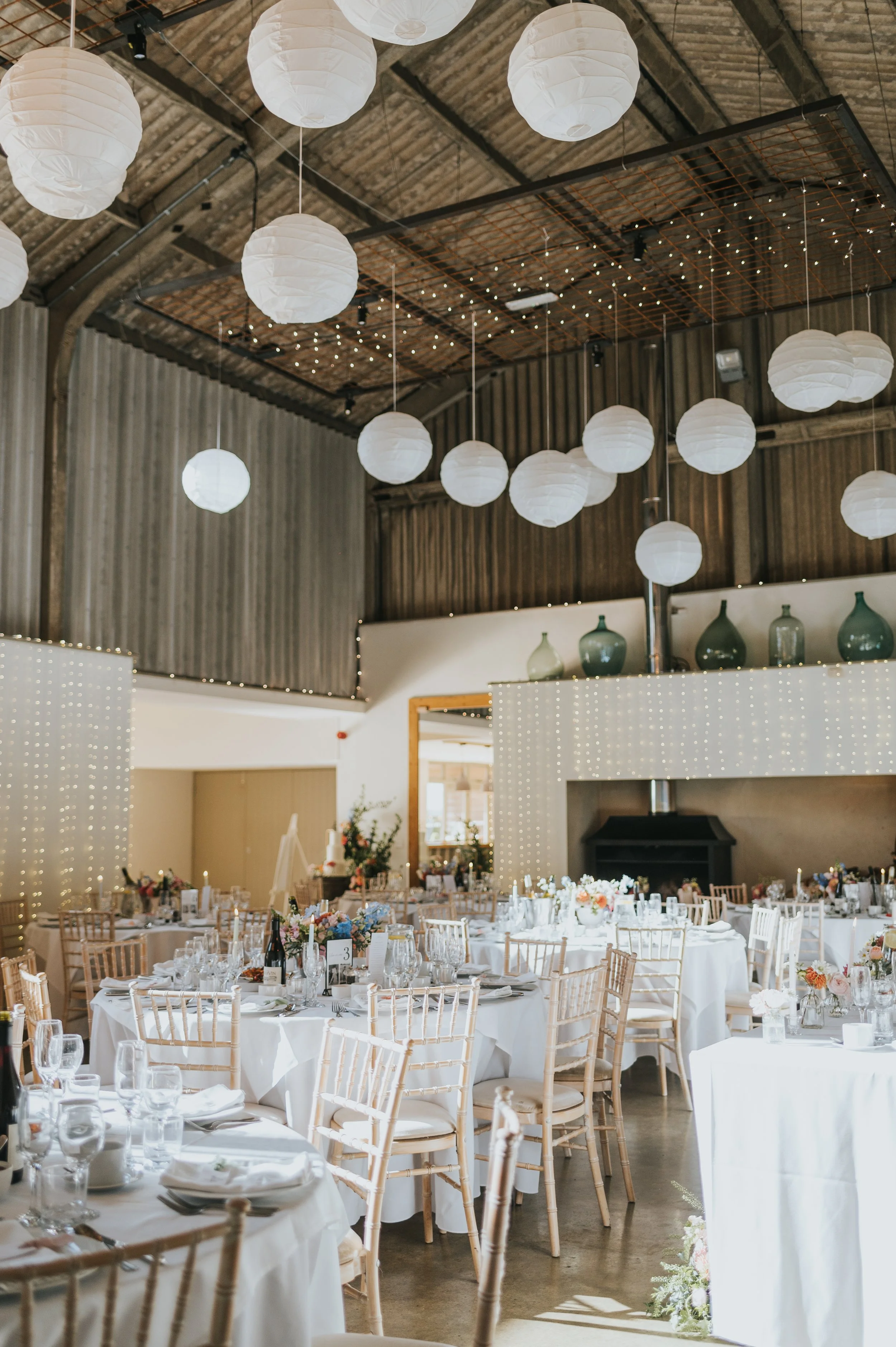 Elegant indoor event space decorated for a celebration with round tables covered in white tablecloths, wooden chairs, floral centerpieces, and string lights, with paper lanterns and vases on a shelf overhead.