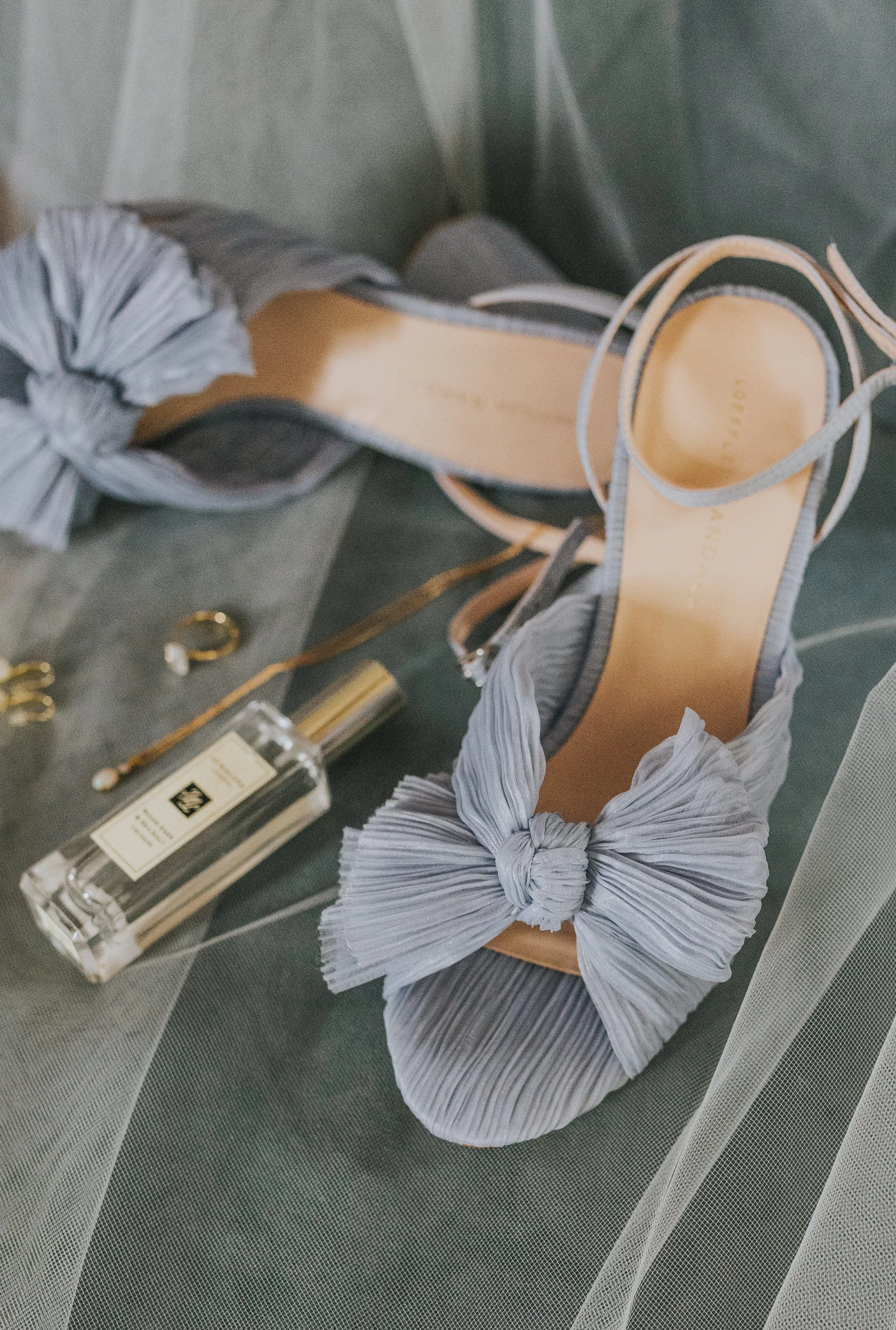 Close-up of a pair of grey high-heeled sandals with pleated fabric bows on the toe straps, positioned on a surface alongside a bottle of perfume and small jewelry items, with sheer fabric draping nearby. Rebecca Louise Photography Essex Wedding photo
