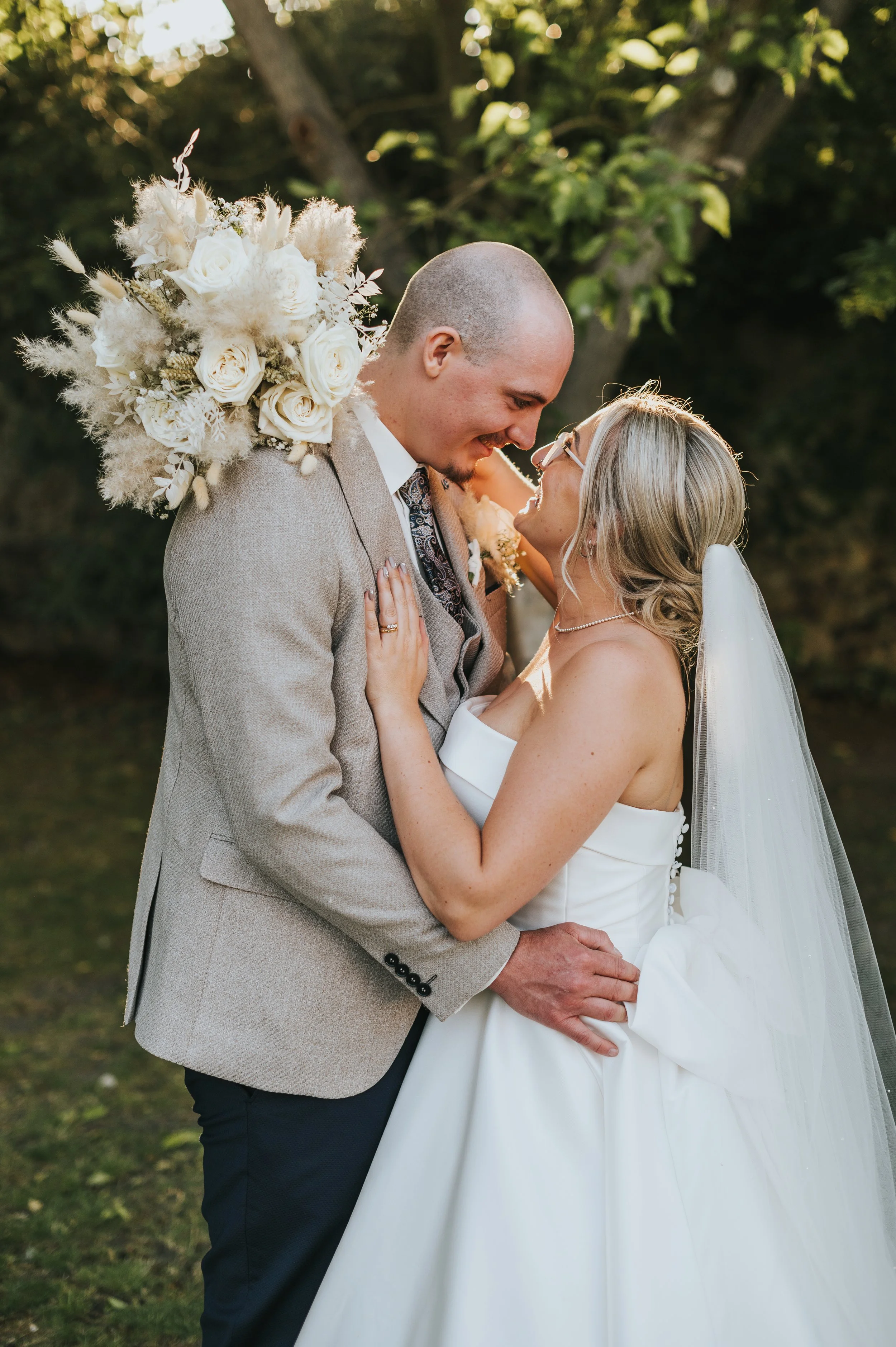 A newlywed couple sharing a joyful moment outdoors, with the bride wearing a strapless white gown and veil, and the groom in a light-colored suit, holding a large bouquet of white and beige flowers.