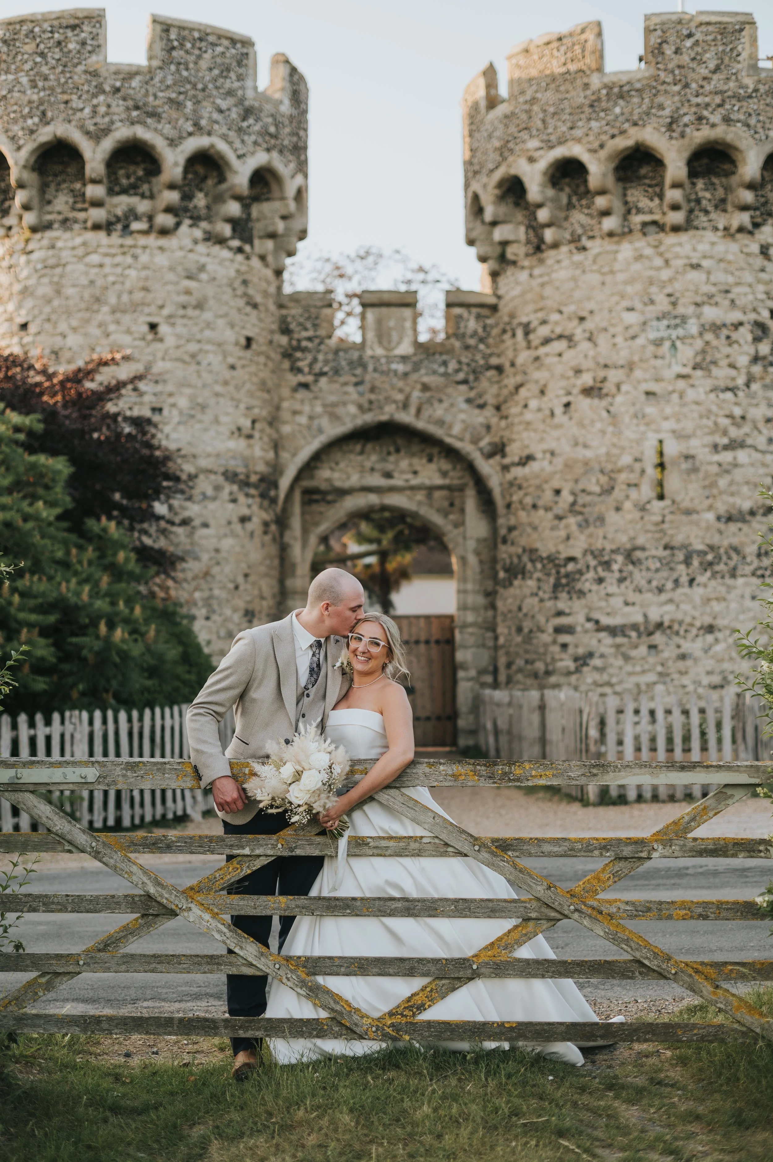A newlywed couple standing behind a rustic wooden gate in front of a medieval stone castle, with the groom kissing the bride's forehead as she smilingly holds a bouquet of white flowers.
