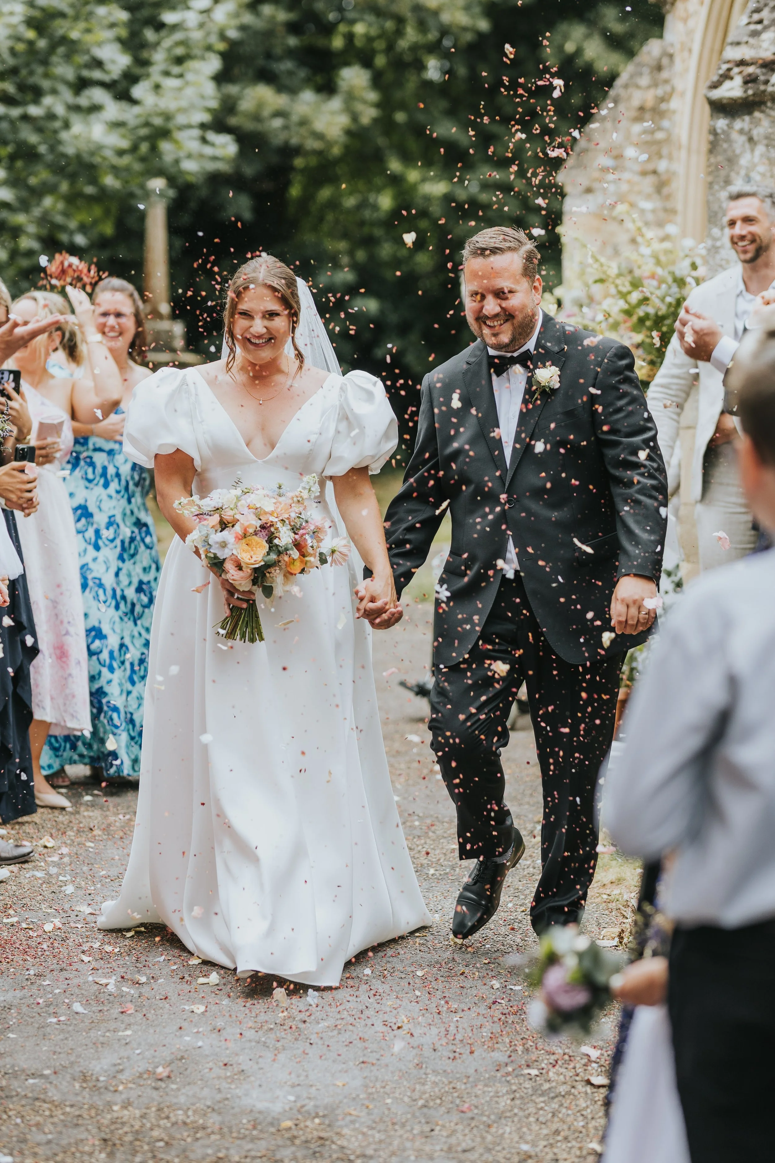 Happy newlywed couple walking hand in hand outdoors while guests celebrate and throw flower petals around them. Rebecca Louise Photography Essex Wedding photographer 
