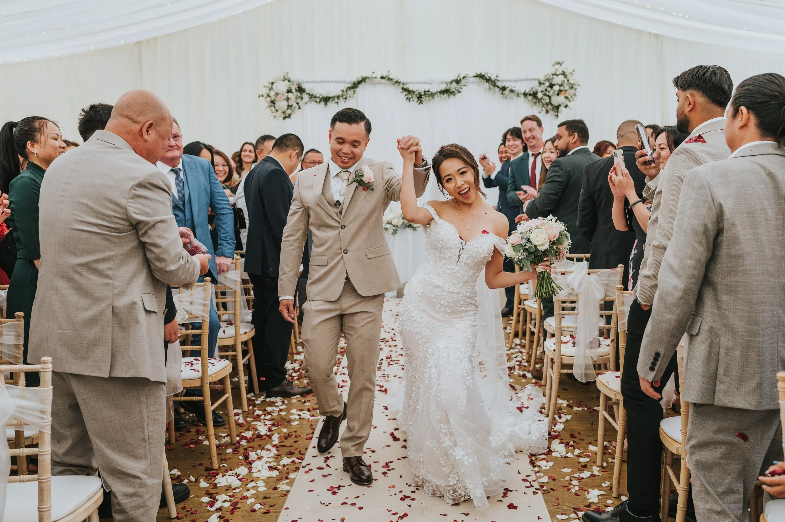 A newly married couple walking down the aisle held by their hands, surrounded by guests in a decorated wedding venue with floral arrangements and chairs, as confetti falls to the ground.