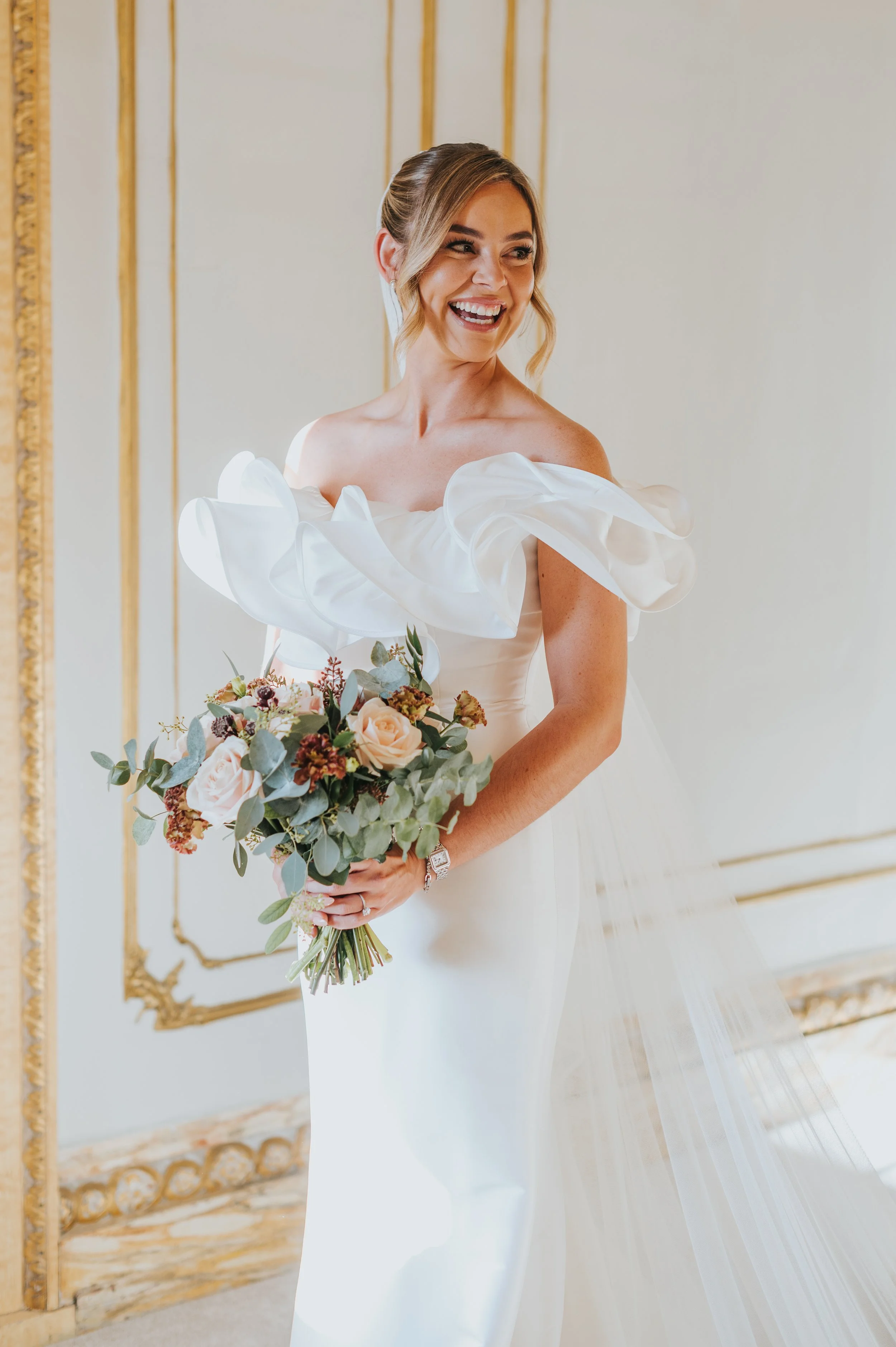A bride in a white wedding dress with ruffled off-shoulder sleeves, holding a bouquet of roses and greenery, smiling in a decorated elegant room.
