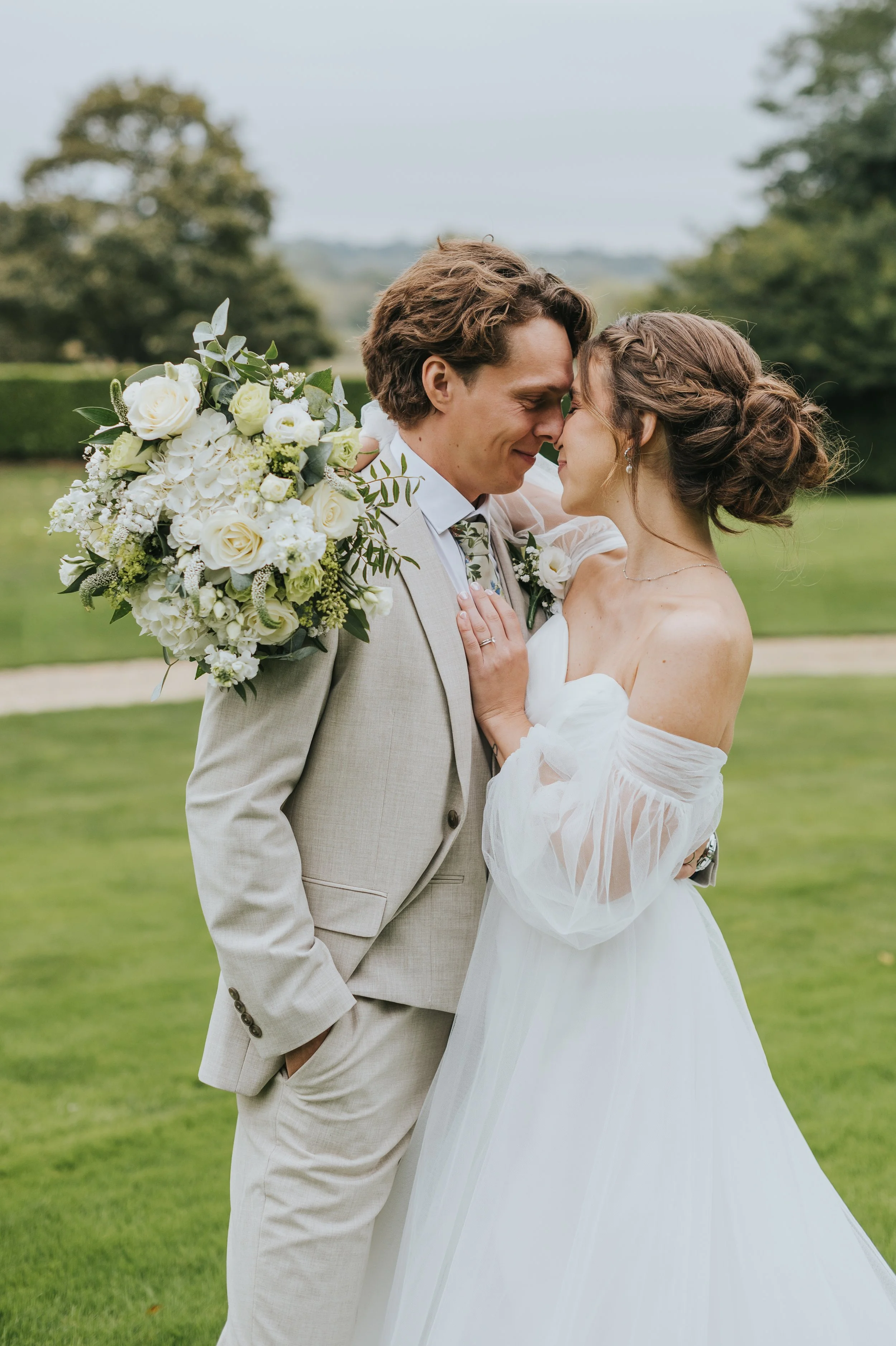 A bride and groom outdoors, touching foreheads, holding a bouquet, smiling, with greenery in the background.