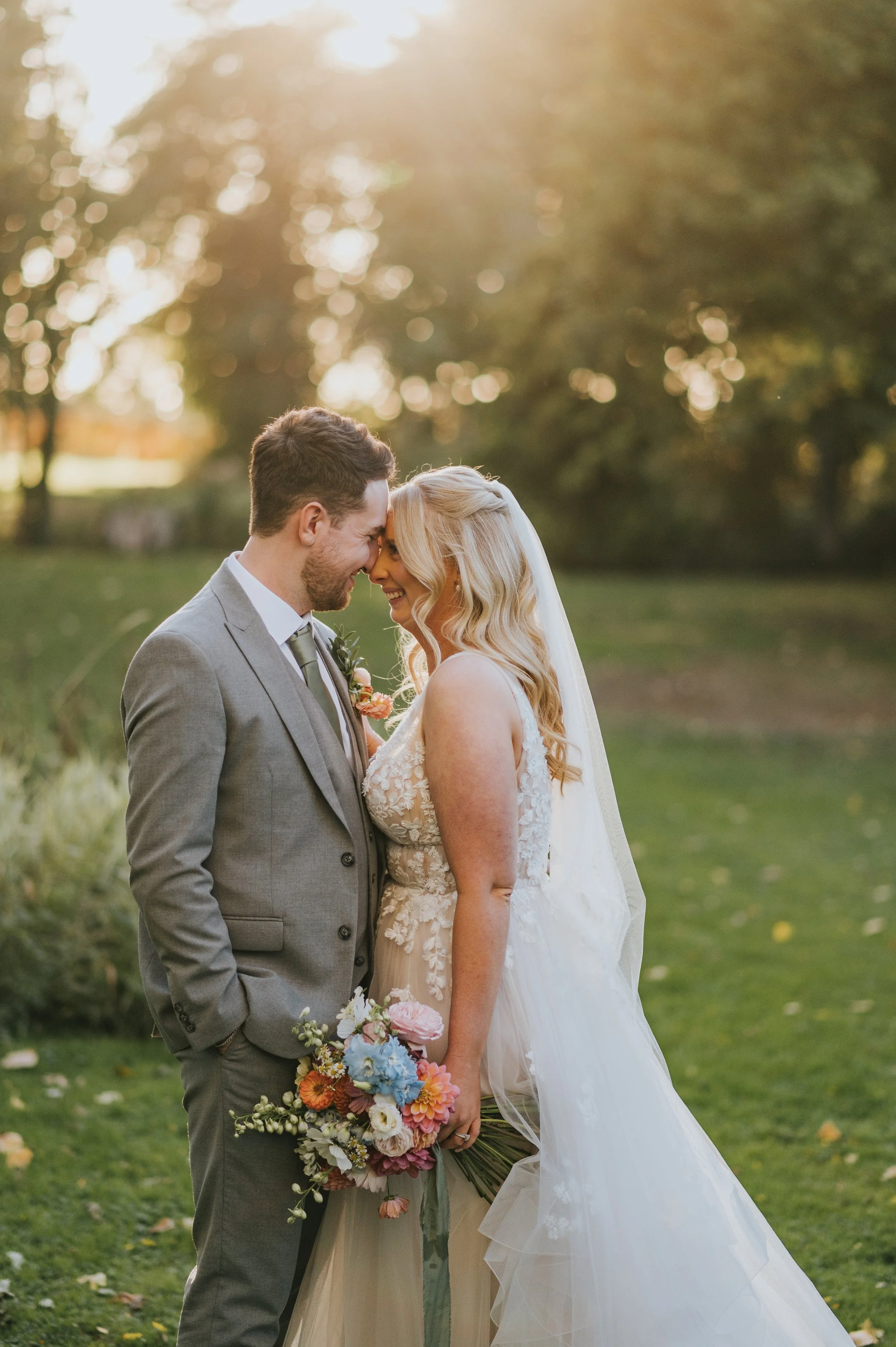 A bride and groom share a kiss outdoors on a wedding day, with sun shining behind trees, holding a bouquet of colorful flowers.