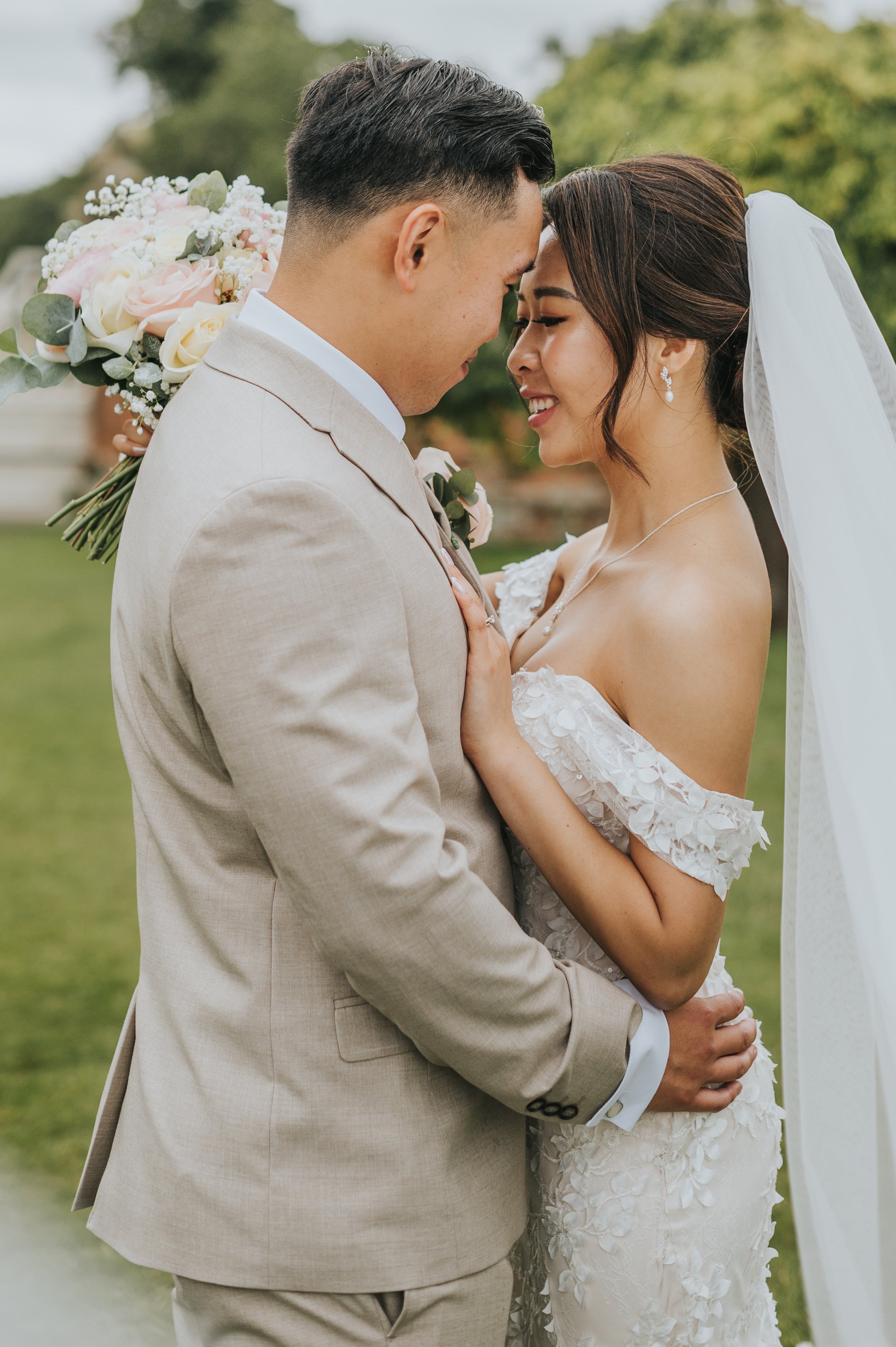 A newlywed couple in wedding attire sharing a close moment outdoors. The groom, wearing a light beige suit, holds the bride, who is in a white wedding gown, and they are touching foreheads with eyes closed, smiling gently.  Quendon Hall wedding