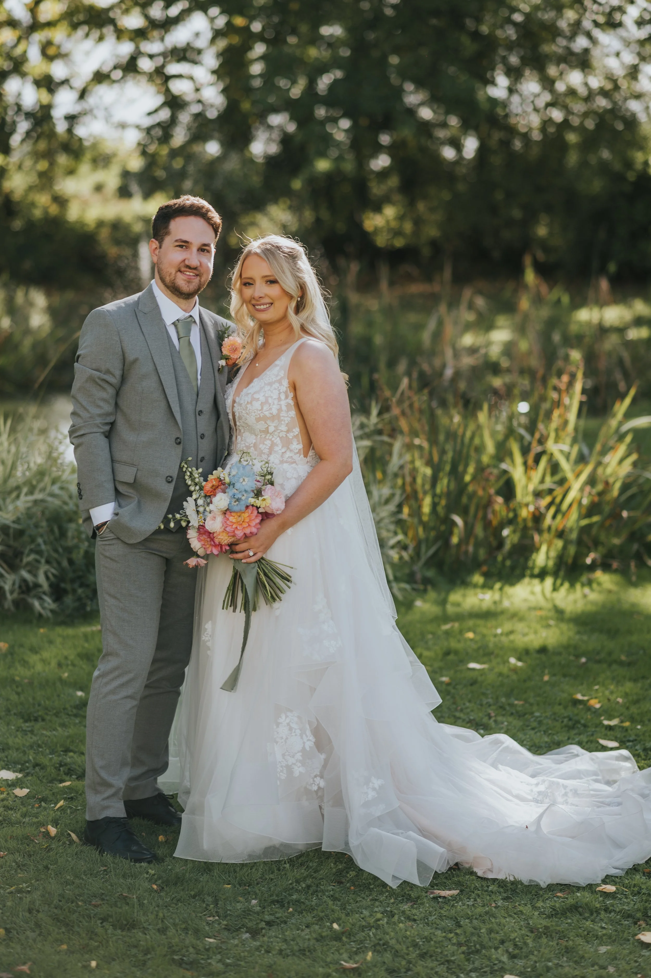 A newlywed couple stands outdoors on a grassy area near a pond, surrounded by trees and foliage. The bride wears a white lace wedding gown with a long train and holds a bouquet of colorful flowers. The groom wears a light gray suit with a white shirt