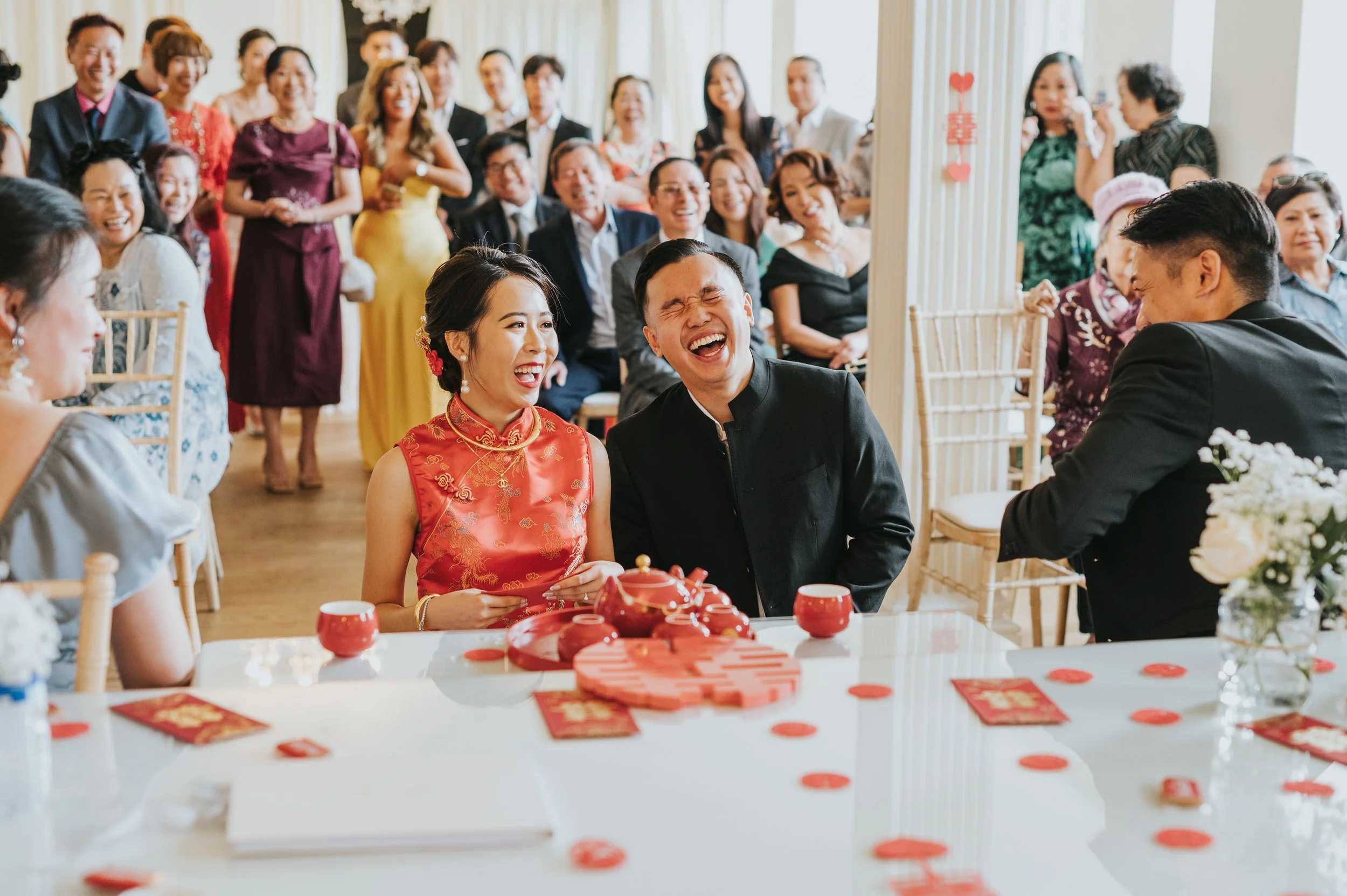 Happy couple at a wedding reception laughing with guests in the background, decorated with red accents
