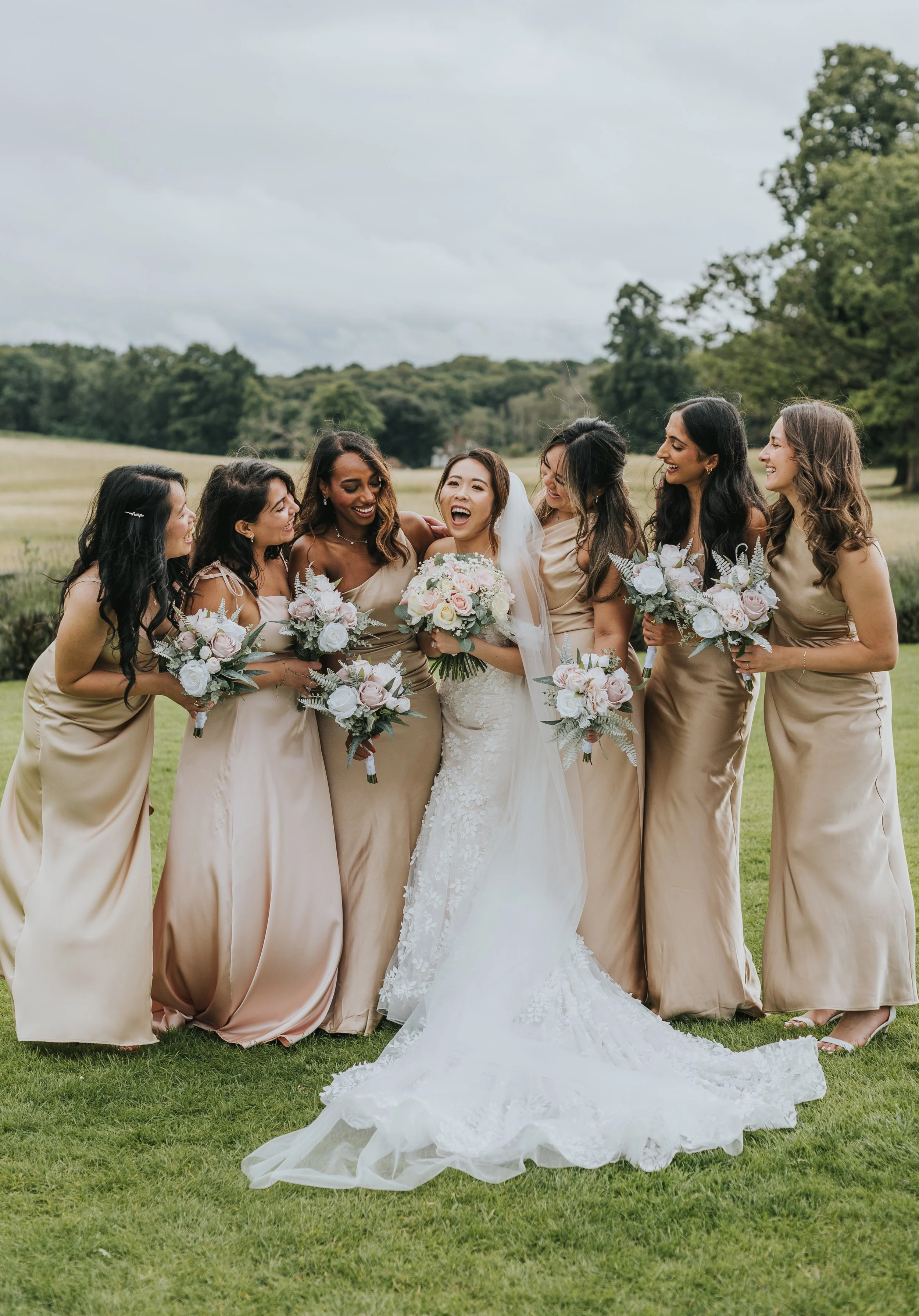 A bride and six bridesmaids standing outdoors on a grassy field, smiling and holding bouquets, with trees and cloudy sky in the background. Rebecca Louise Photography Essex Wedding photographer Quendon Hall