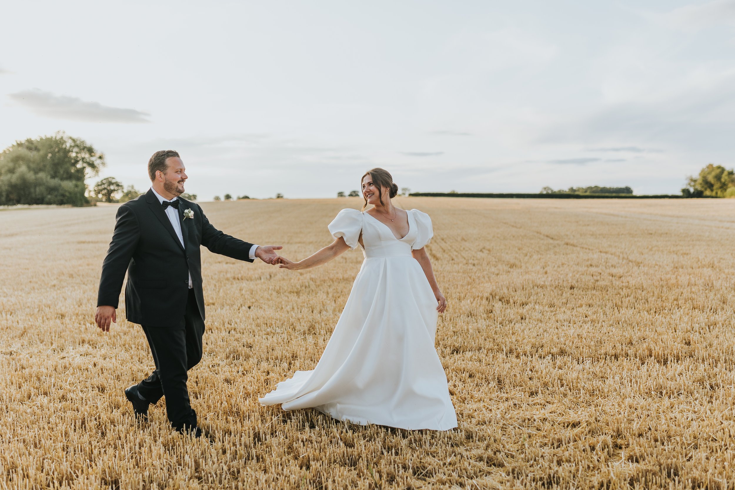 A bride and groom holding hands and smiling in a wheat field during their wedding.