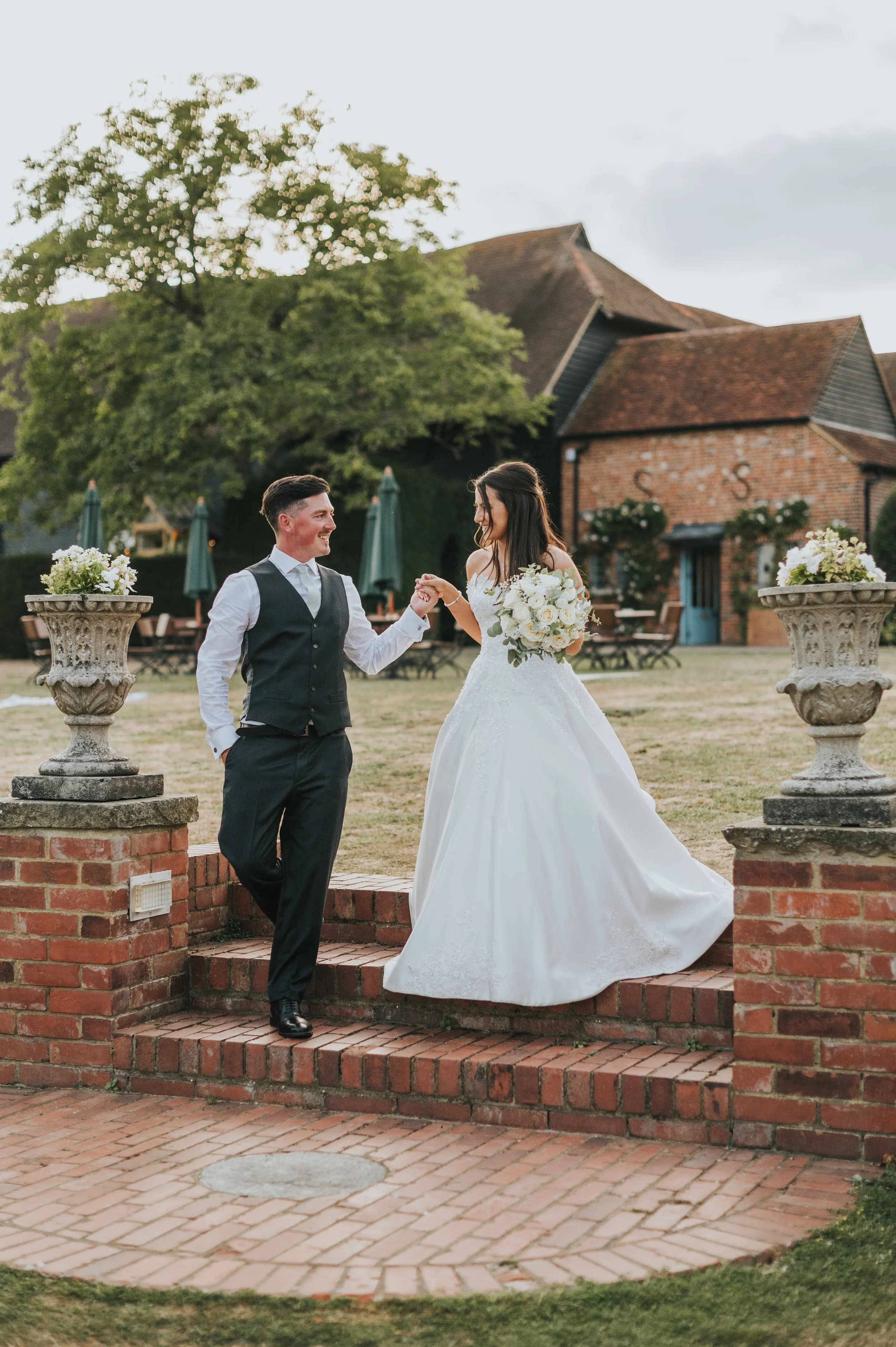 A bride and groom standing on steps outdoors, holding hands and smiling at each other, with wedding flowers and a rustic building in the background.