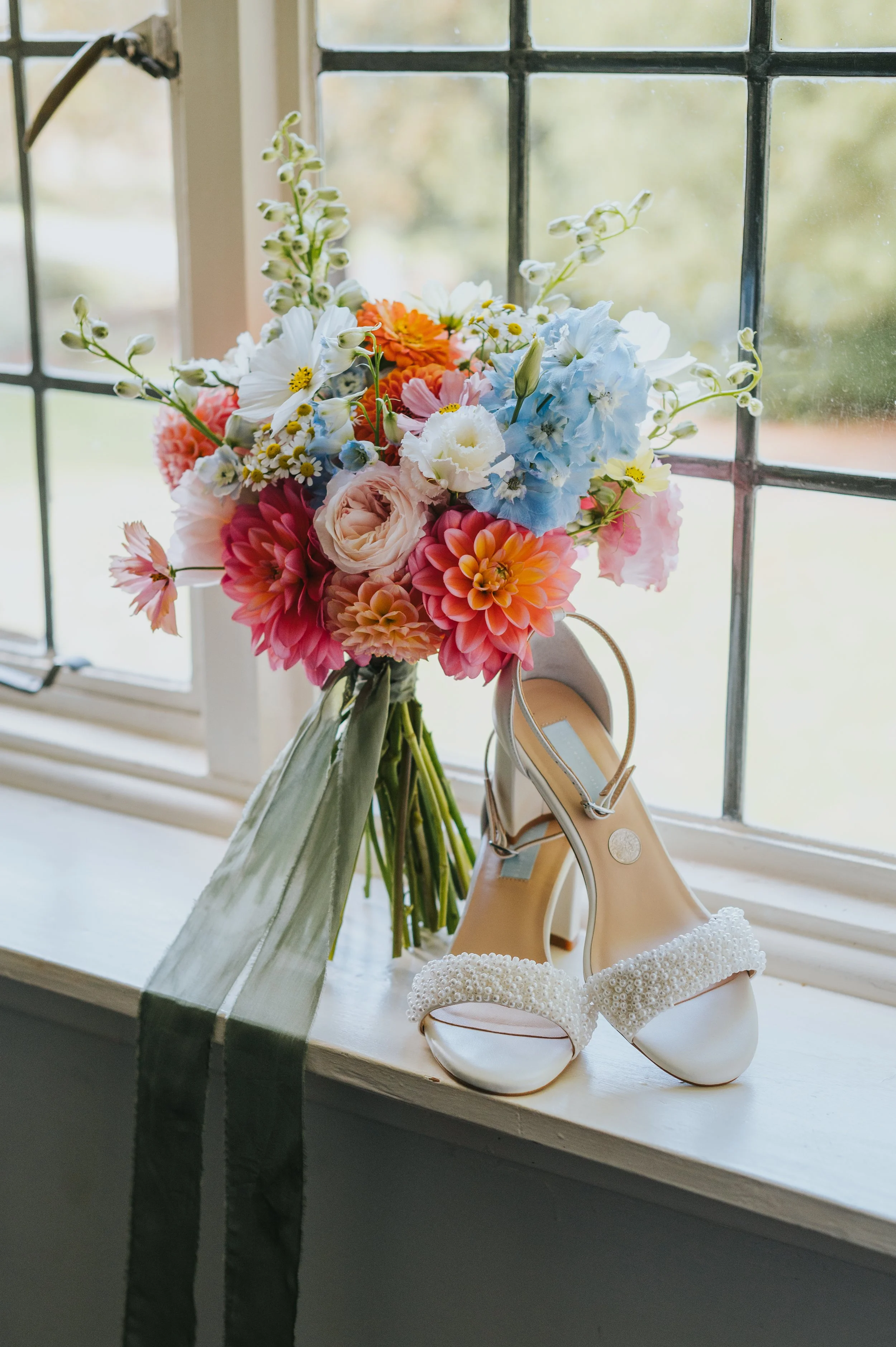 A bouquet of colorful flowers and a pair of white wedding shoes on a window sill.