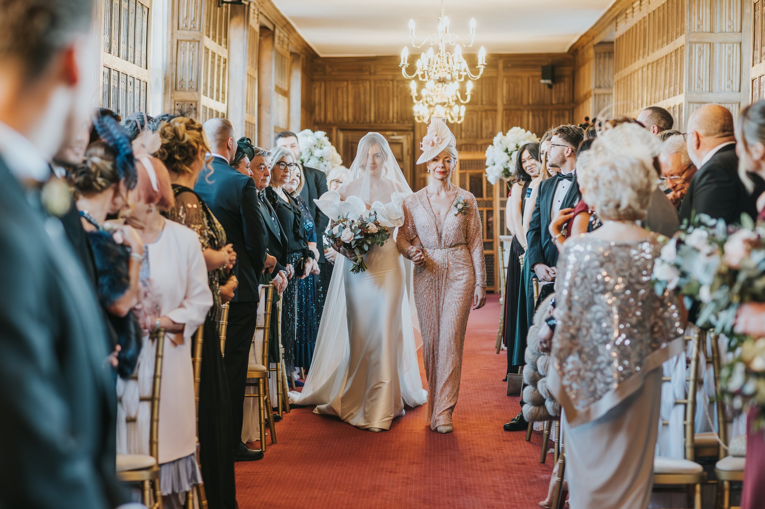 A bride in a wedding gown walking down the aisle, accompanied by an older woman in a rose gold sequin dress, in a decorated wooden banquet hall with chandeliers and floral arrangements, surrounded by seated guests.