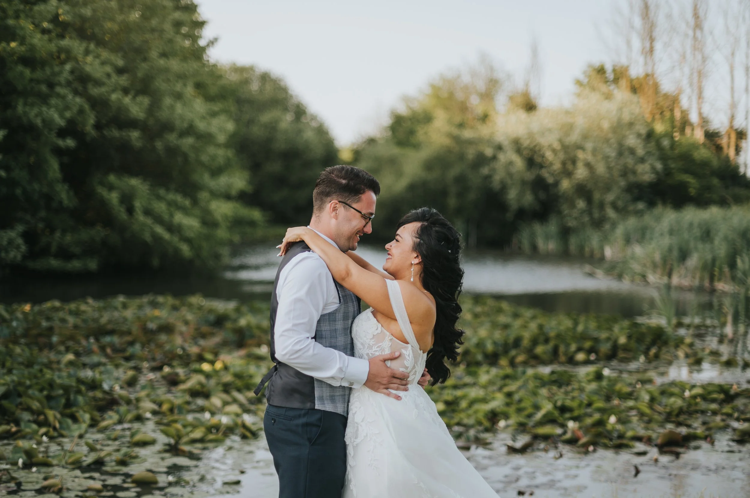 A couple dressed in wedding attire embracing near a pond surrounded by greenery. Rebecca Louise Photography Essex Wedding photographer