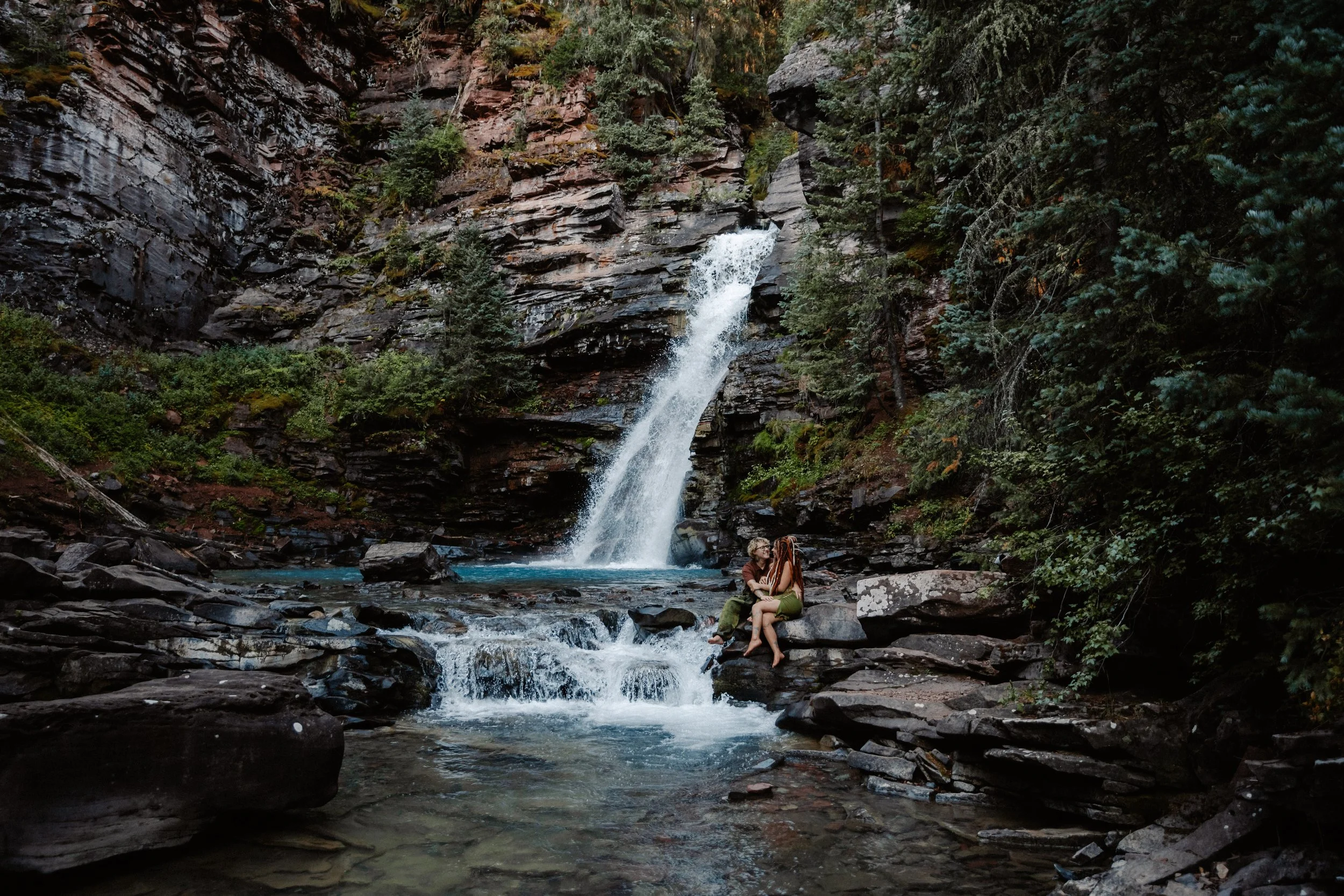 A couple sitting on rocks near a waterfall in a forested canyon