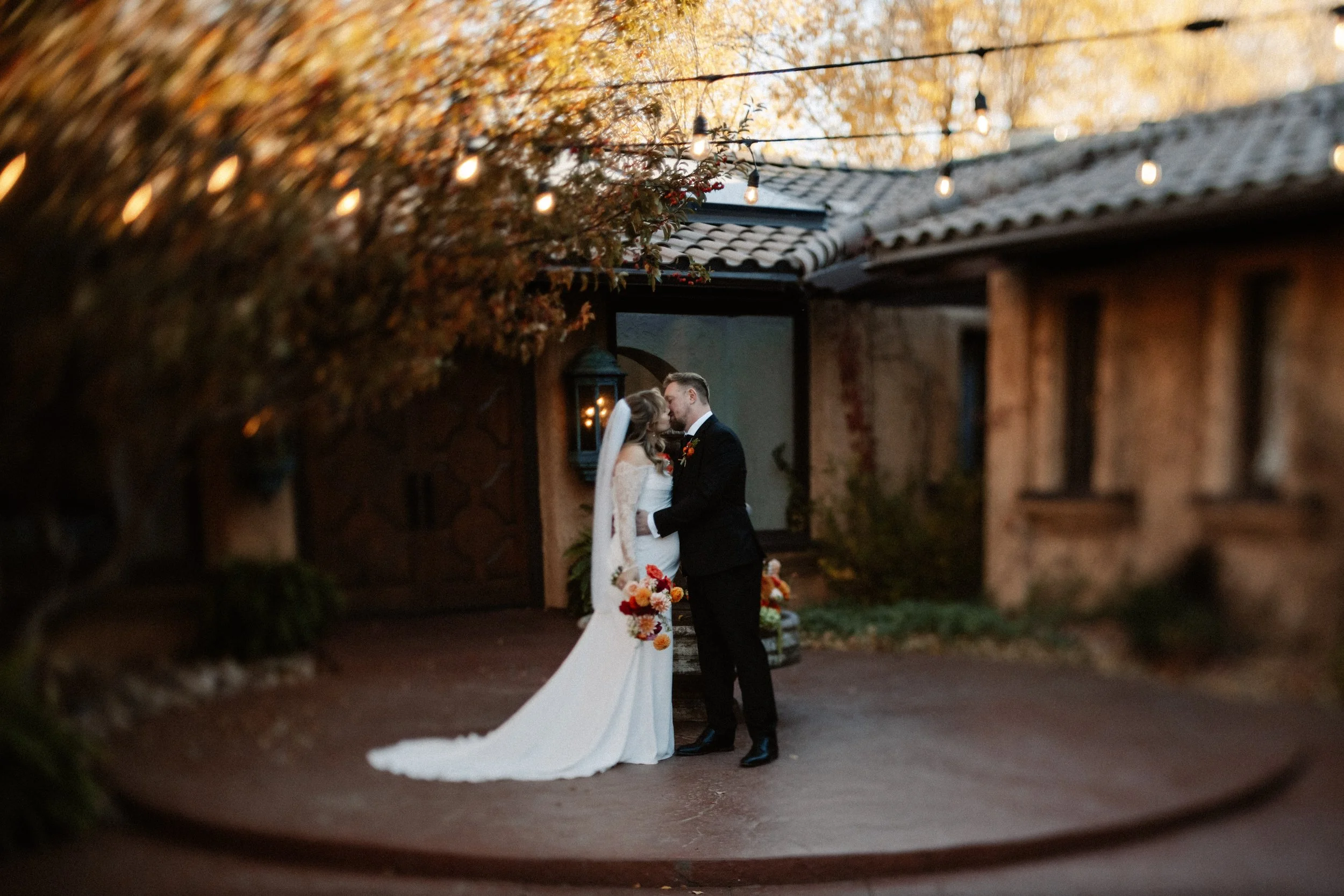 A bride and groom sharing a kiss outdoors during their wedding, with warm lighting and a rustic building in the background.