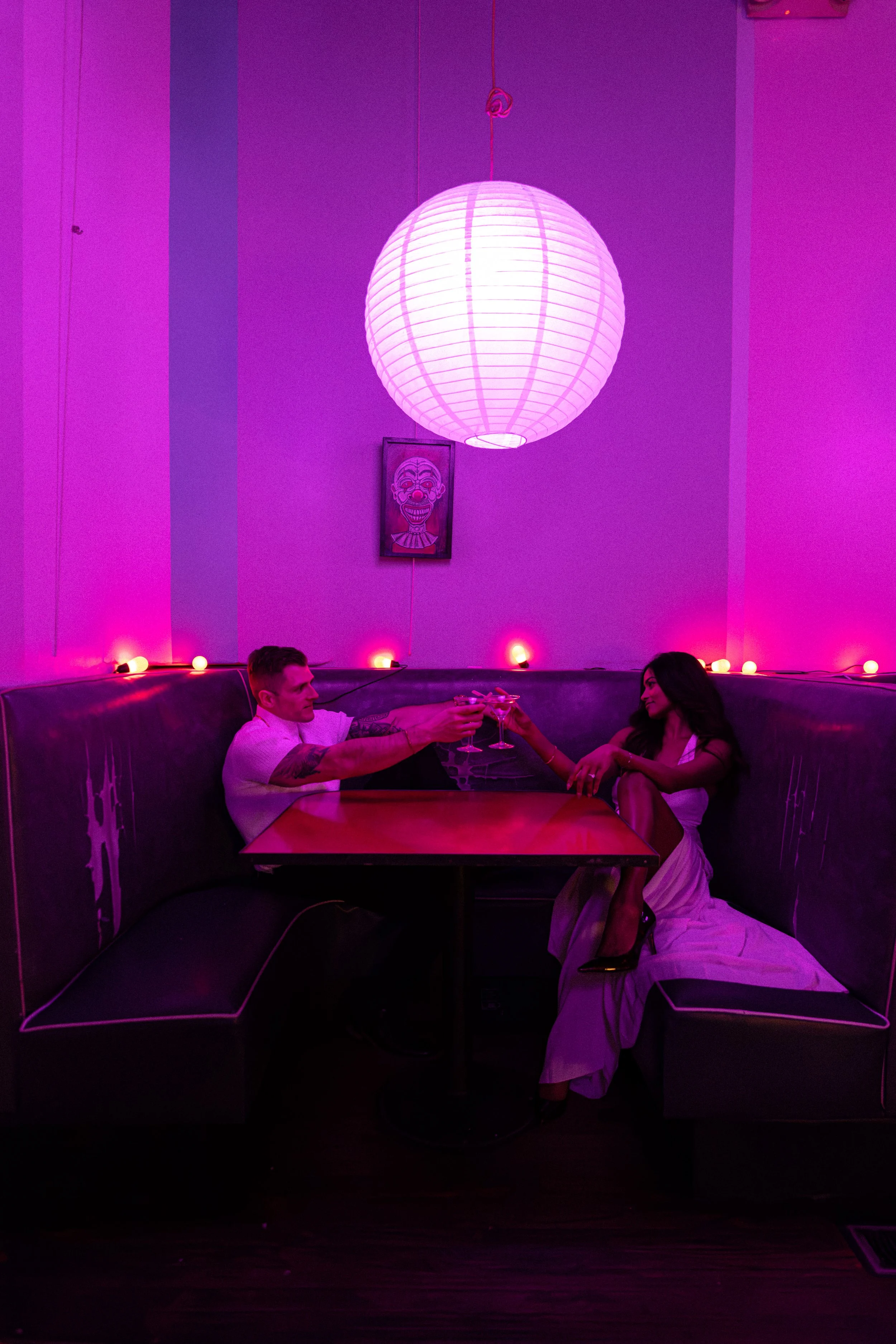 A man and woman sitting in a booth, clinking glasses under a large pink paper lantern light, with purple and pink lighting creating a vibrant atmosphere.