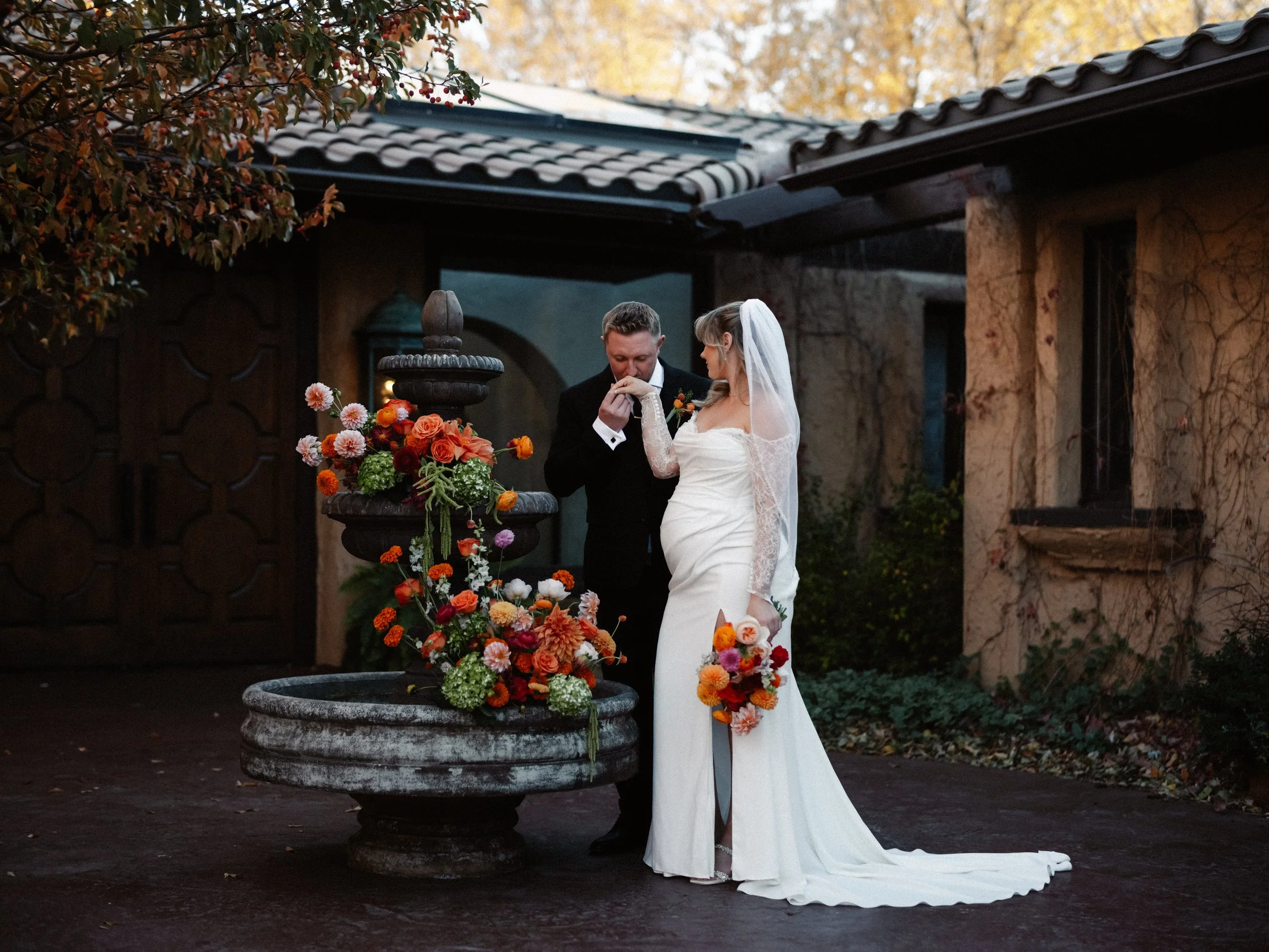 A bride and groom stand by a fountain decorated with colorful flowers during their wedding ceremony outdoors.