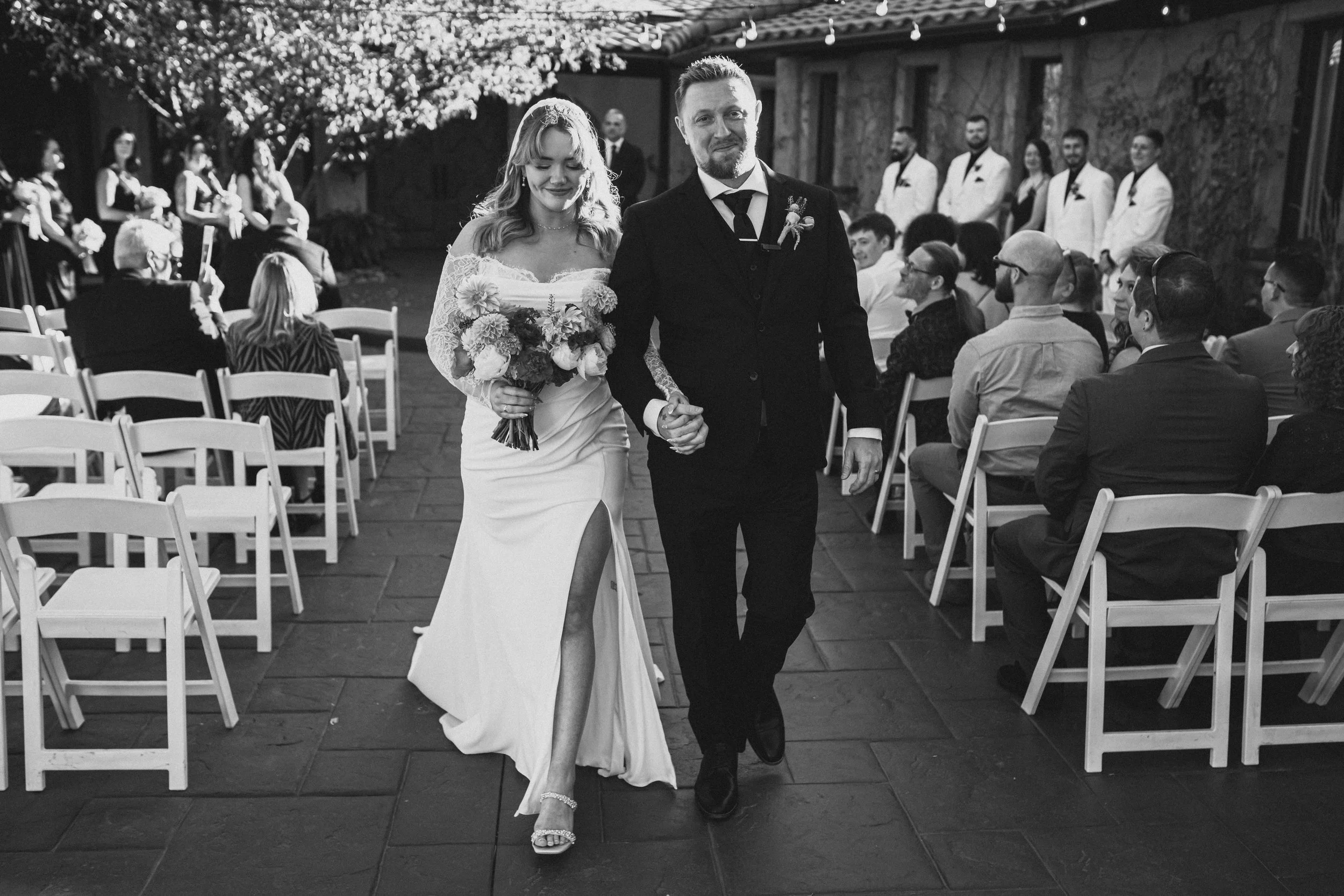 A bride and groom walking down the aisle at their outdoor wedding ceremony surrounded by seated guests and groomsmen in white tuxedos.