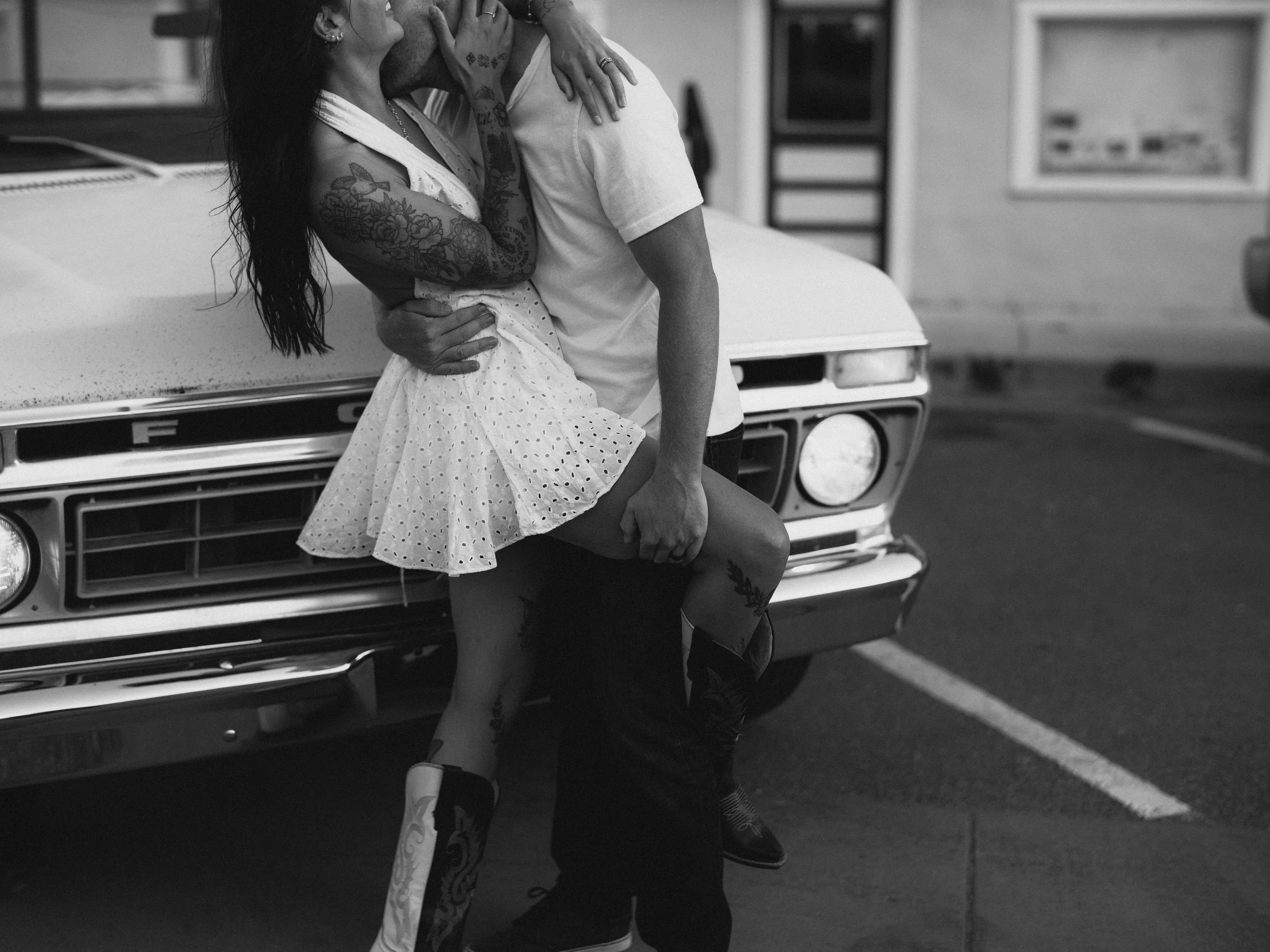 A woman and man kissing in front of a vintage car in a parking lot, with the woman's legs and tattoos visible, black and white photo.