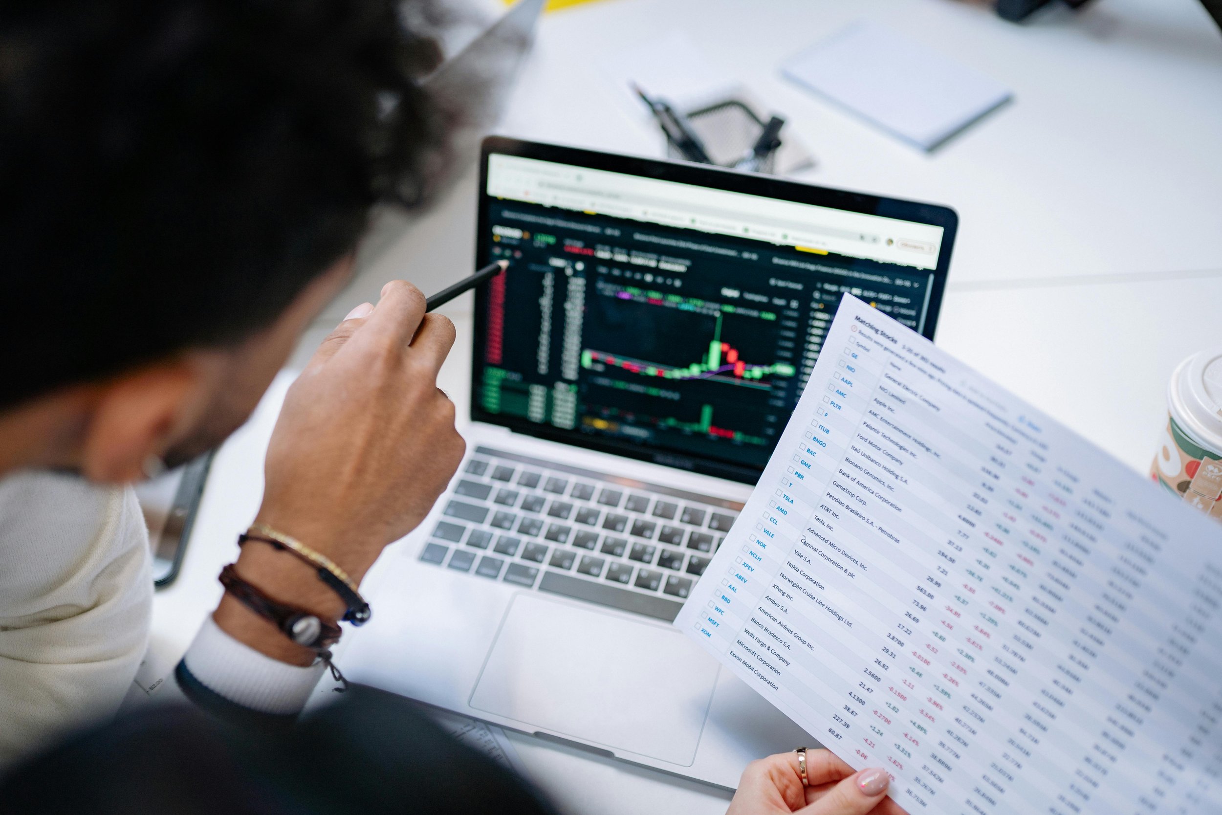 Person analyzing stock market data and technical chart on laptop with a printed financial report, coffee cup, and notepad on table.