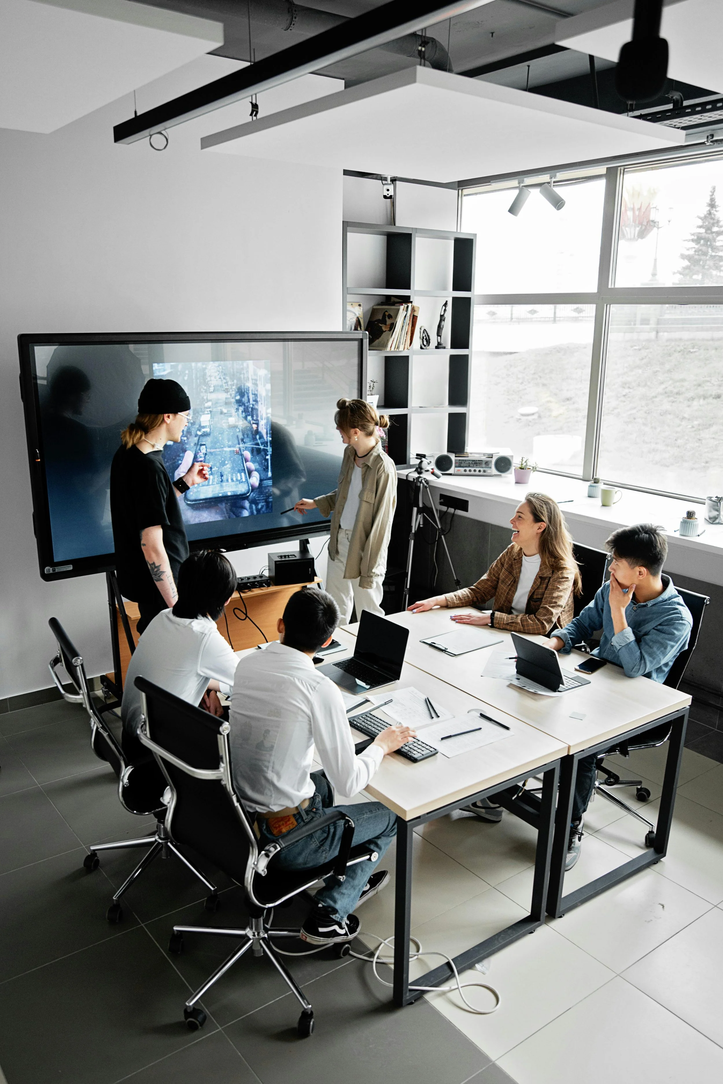 A group of five young adults in a modern conference room with a large screen displaying a cityscape, engaging in a presentation or discussion, with laptops and notebooks on the table and a large window providing natural light.