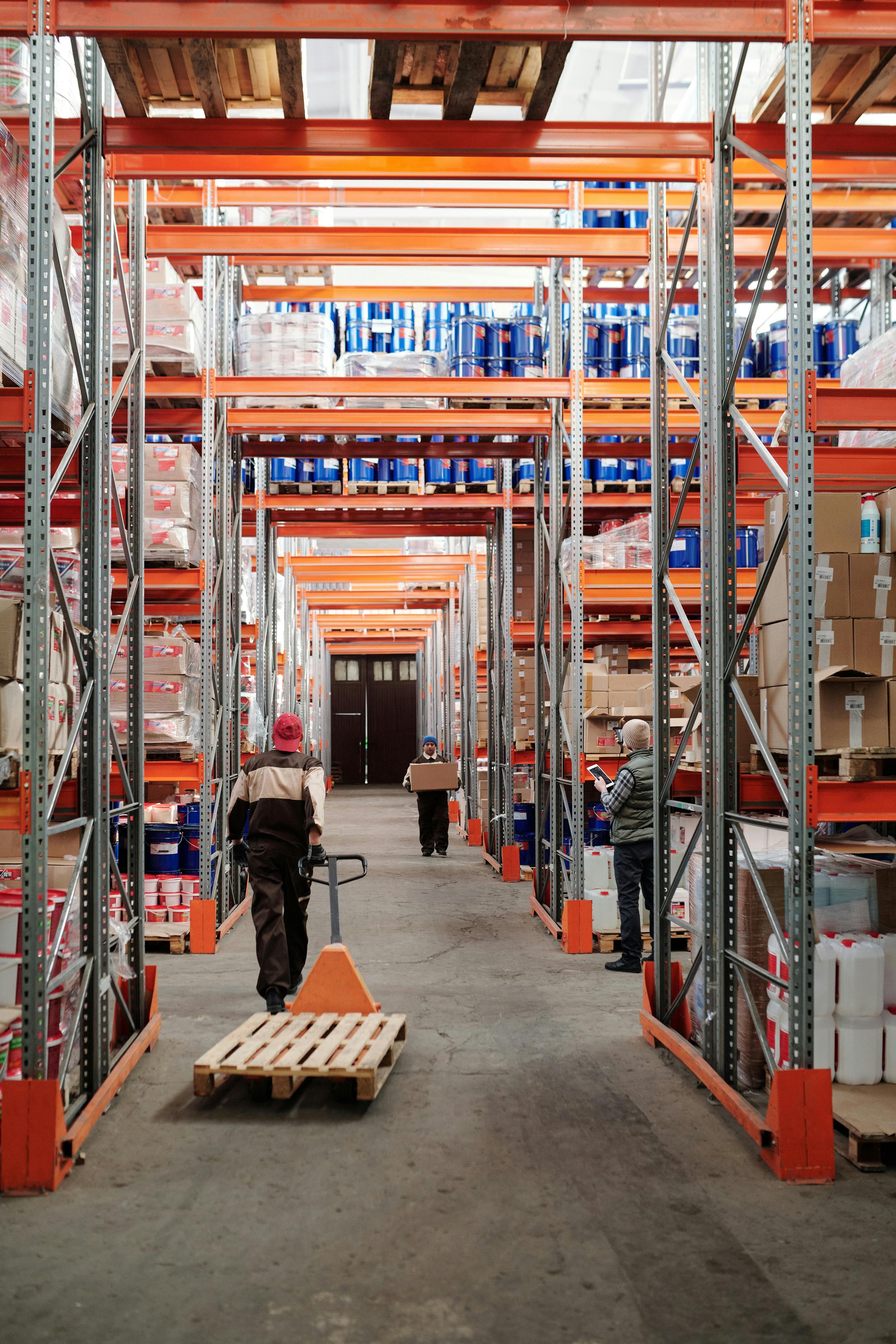 Warehouse scene with tall metal shelves stacked with boxes and cans, three workers organizing stock, one using a pallet jack, another holding a box, and a third looking at a tablet.