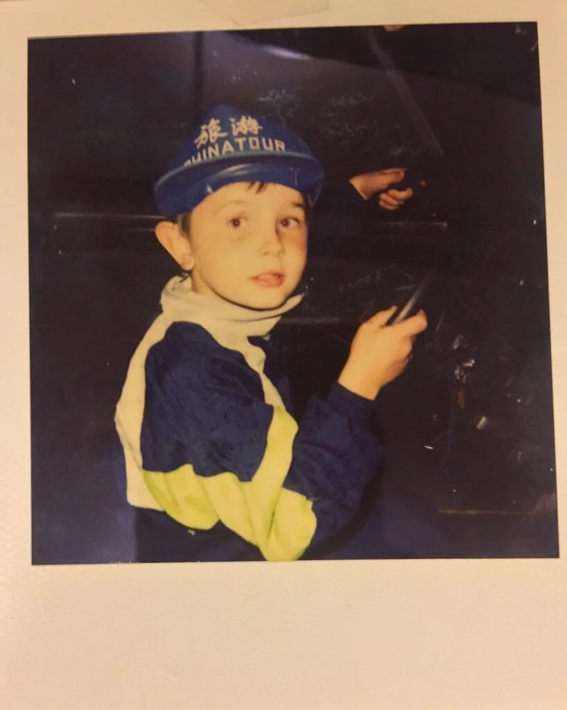 Young boy wearing a blue helmet labeled 'FUN AT TOUR', sitting in a vehicle, holding a steering wheel, looking at the camera.