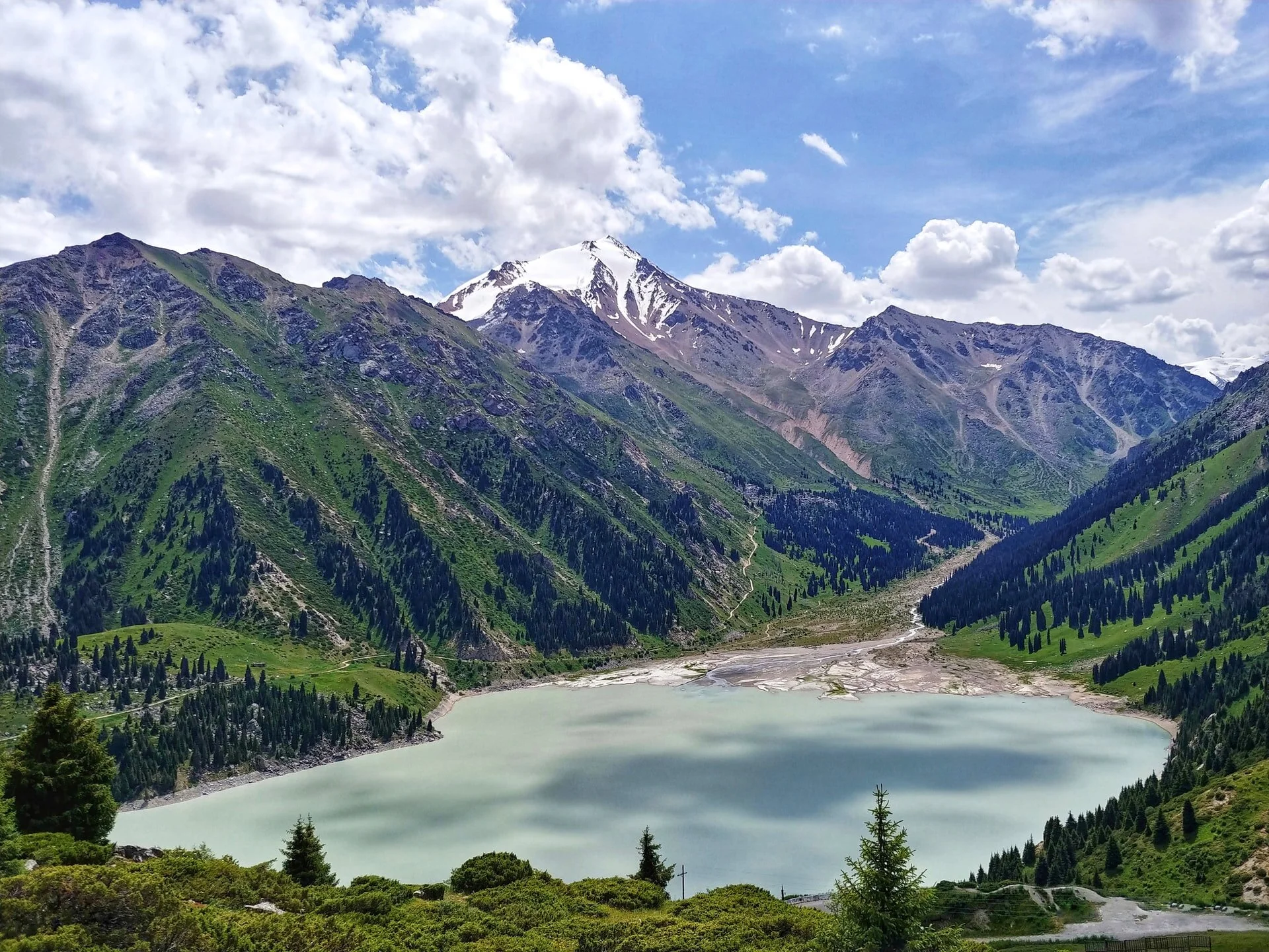 Snow-capped mountain above green forested hills and a glacial lake in a mountainous landscape.
