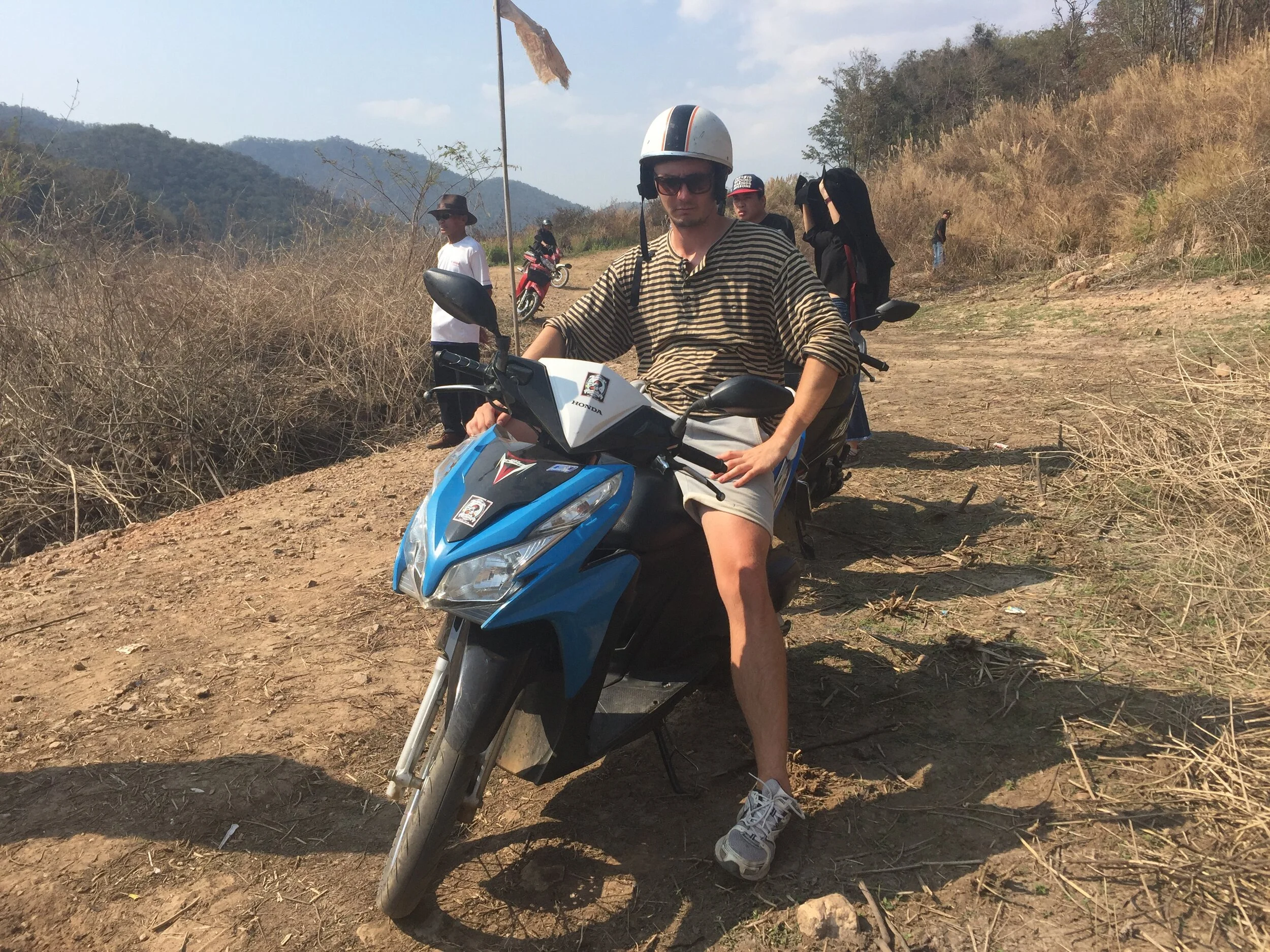 A man wearing a helmet, sunglasses, a striped shirt, and shorts sitting on a blue motorbike on a dirt trail in a dry, hilly outdoor area with a few people and motorcycles in the background.