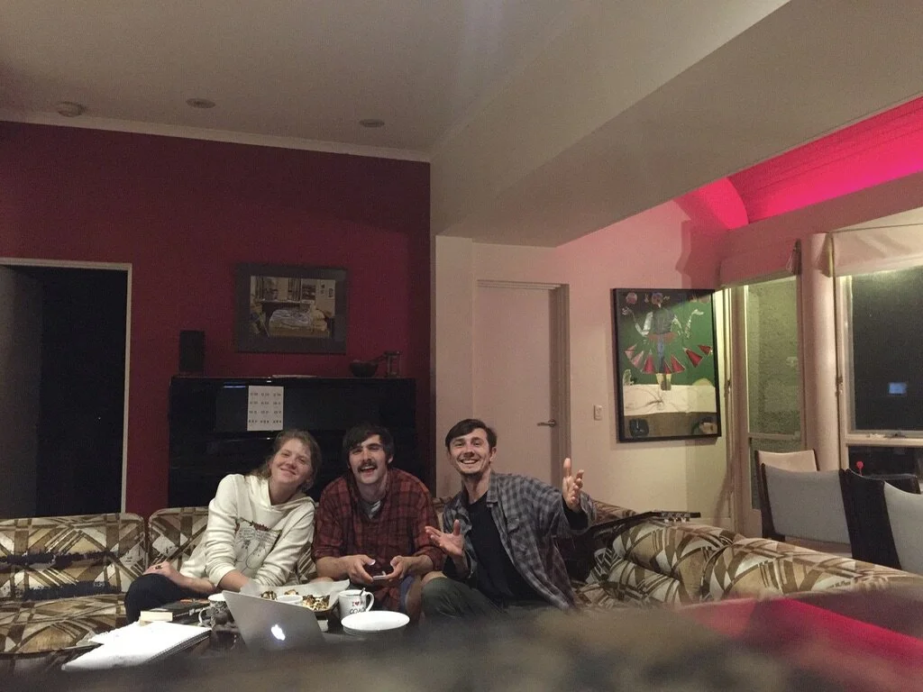 Three young adults sitting on a patterned sectional sofa in a living room, smiling and enjoying each other's company. There are papers, a laptop, and dishes on the table in front of them, with a piano and colorful artwork visible in the background.